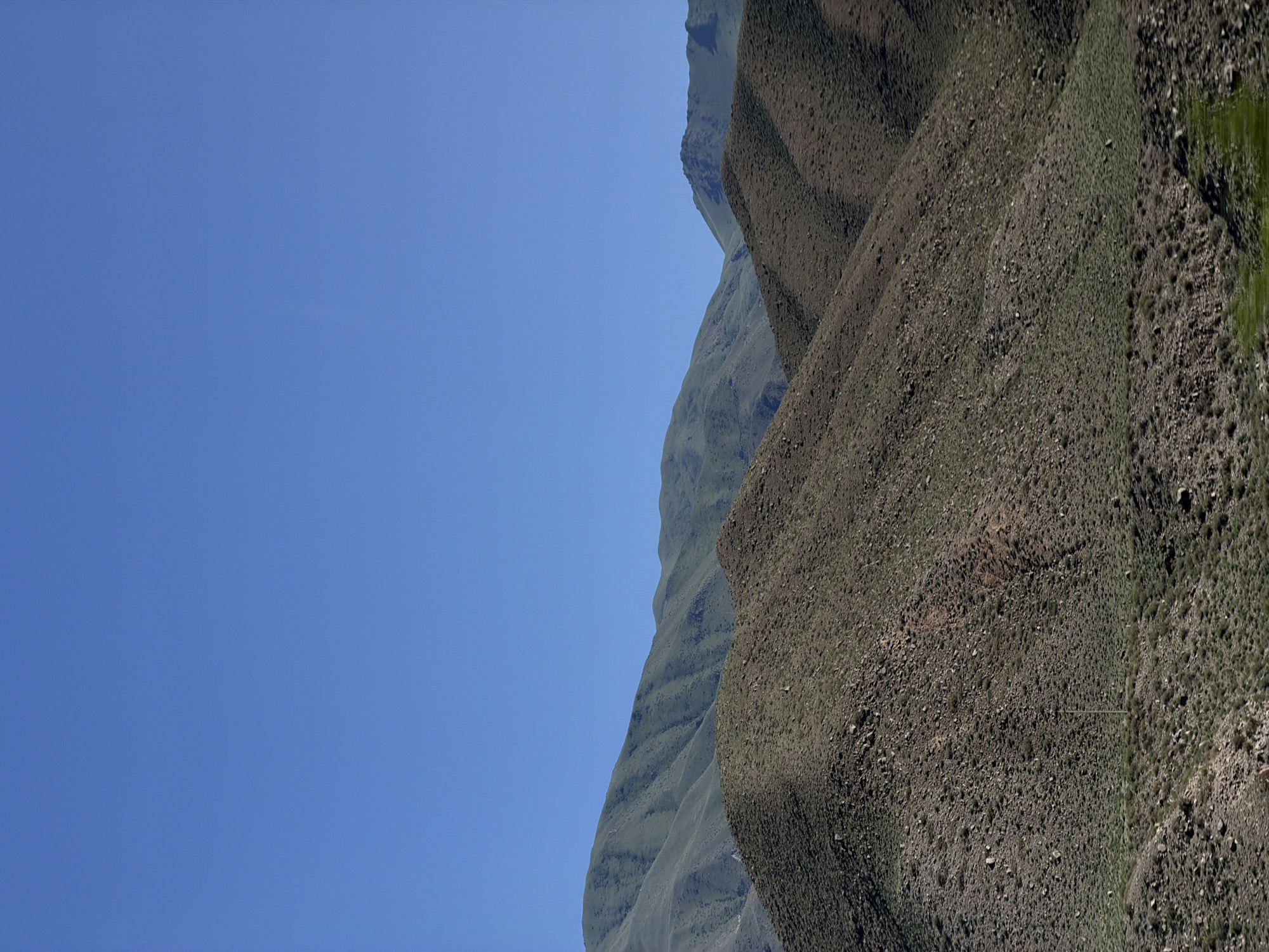 The image displays a vast, clear blue sky on the left, filling roughly two-thirds of the frame. On the right, a steep, rugged mountain slope dominates the scene. The mountain is covered in sparse vegetation and rocks, with visible texture and varied shades of brown and green. The terrain appears arid and uneven, with shadows indicating the contours of the landscape. The sunlight suggests it is daytime. The overall impression is one of natural, remote wilderness, likely in a mountainous region. There are no people, objects, or visible text in the image. The location context provided is Kok Moynok Eki, Kyrgyzstan.
