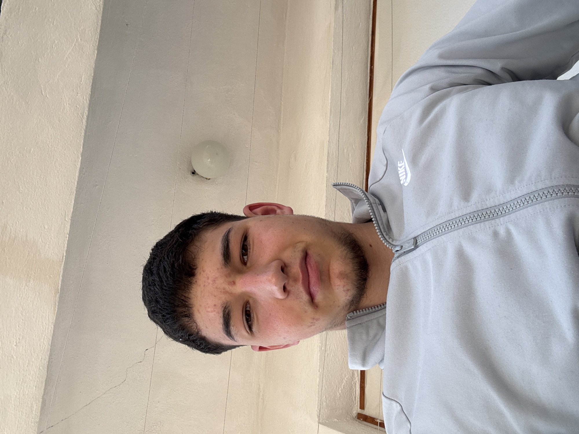 A young man is posing for a selfie indoors. He has dark hair and brown eyes and is wearing a light gray Nike tracksuit. The background shows a white ceiling with a simple light fixture. The location in Zlatograd, Bulgaria, cannot be determined from this image alone.