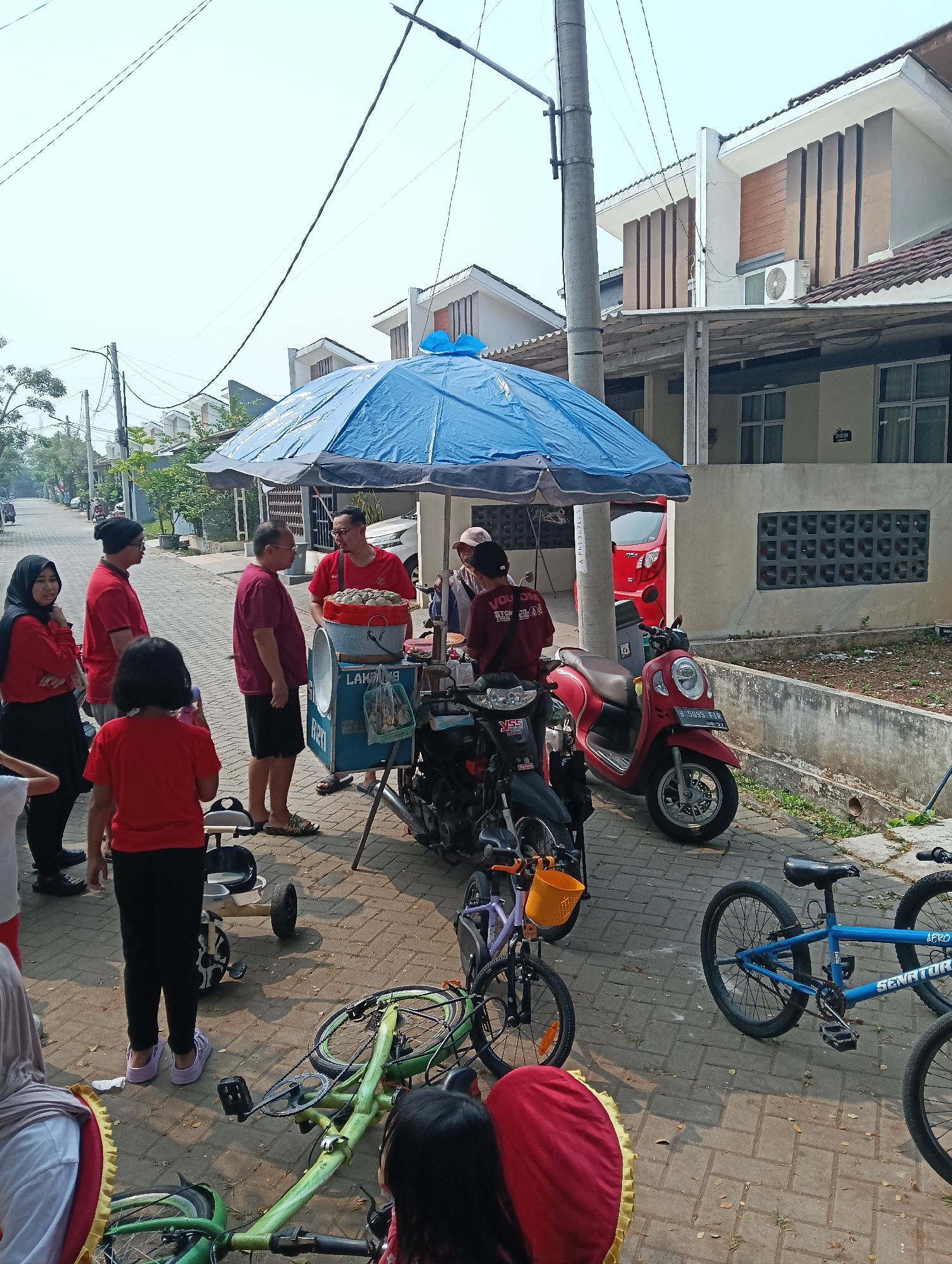 The image captures a street scene in Parung Panjang, Indonesia, likely during daylight hours due to the bright sunlight. A group of people, some wearing red shirts, are gathered around a food cart parked under a large blue umbrella. The cart appears to be selling some kind of food, possibly meatballs, served from a large red bowl. There are motorcycles parked nearby, including a red scooter. In the foreground, children's bicycles, one green and one purple, are visible, one of them lying on its side. Houses with modern architecture line the street in the background. The overall atmosphere suggests a casual outdoor gathering or street market.
