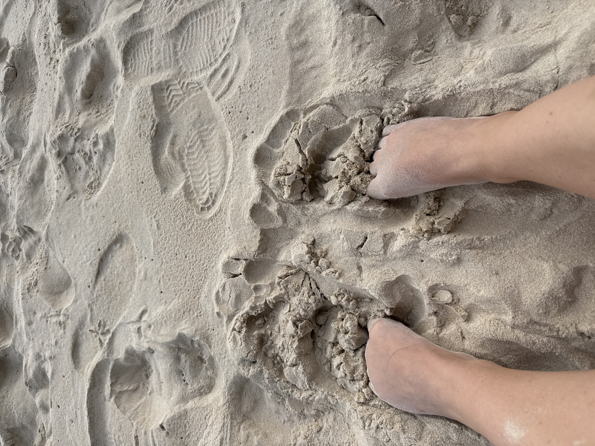 The image shows a close-up, overhead view of two bare feet standing in light brown sand. The feet are partially buried in mounds of sand, with toes visible and dusted with more sand. The surrounding sand is textured, showing numerous footprints and indentations from what appear to be both human and bird tracks. The lighting suggests it is daytime. The location is Karon Beach, Karon, Thailand.