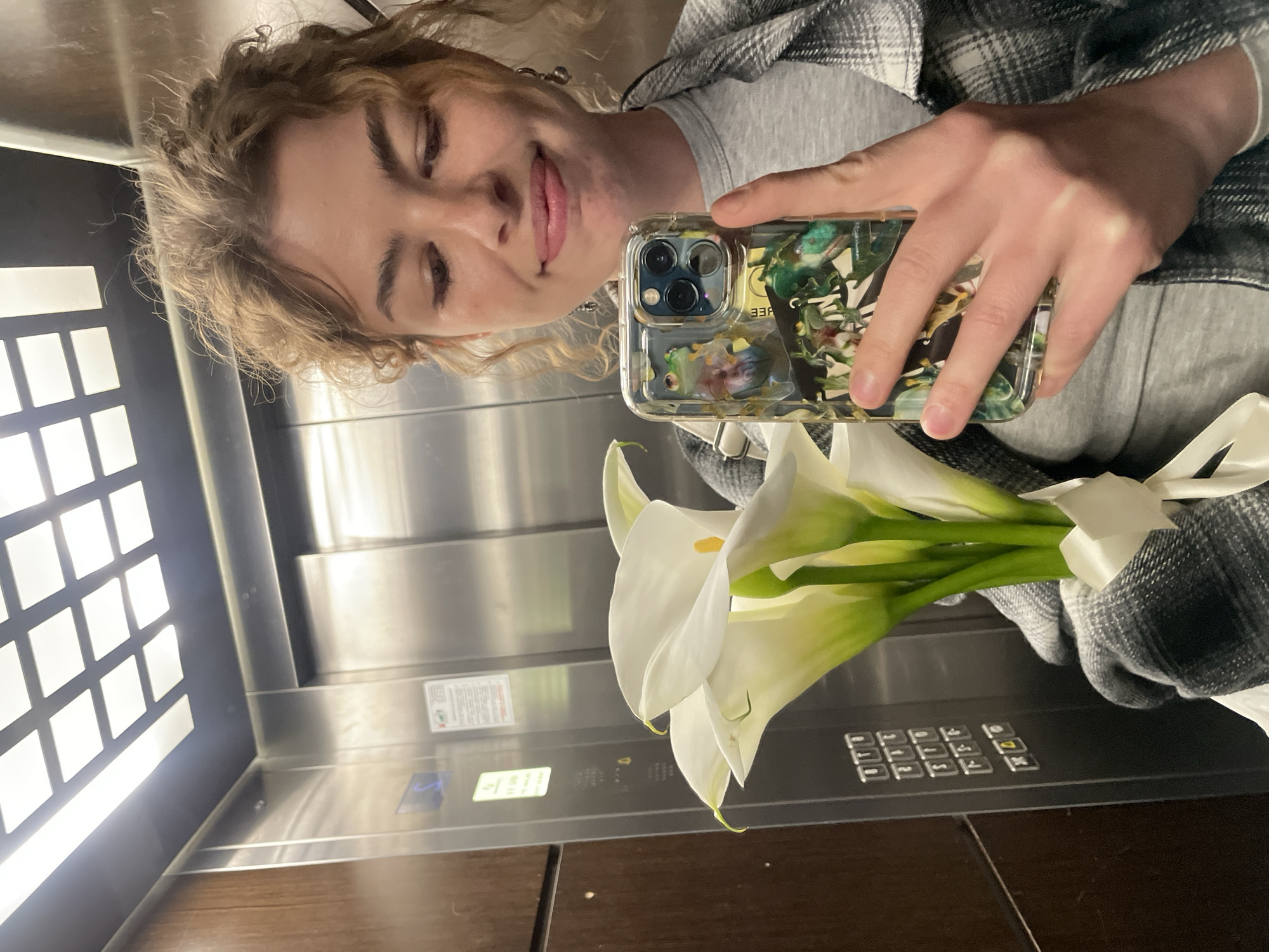 A young, light-skinned woman with curly blonde hair is taking a selfie in what appears to be an elevator. She is holding a bouquet of white calla lilies in front of her. She is wearing a light grey t-shirt and a grey and black plaid shirt over it. The woman is holding a cell phone with a clear case decorated with what seems to be frogs. The background shows the elevator interior, including the panel of buttons and the textured wall. The location in Boreti, Montenegro, is not discernible.