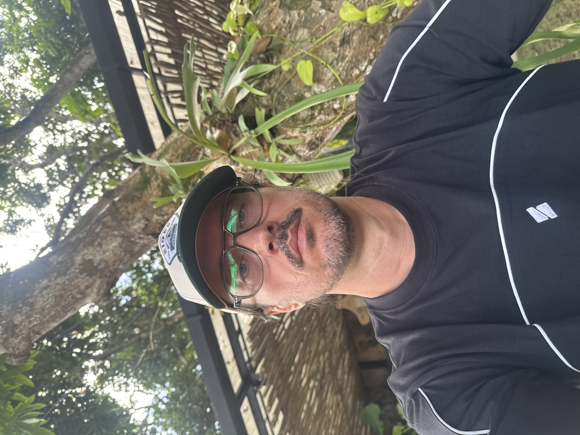 A middle-aged man with glasses and a baseball cap is posing for a selfie outdoors. He is wearing a dark-colored athletic shirt with white trim. The background includes a tree, plants, and a wooden structure. The image was taken in Kuta Selatan, Indonesia.