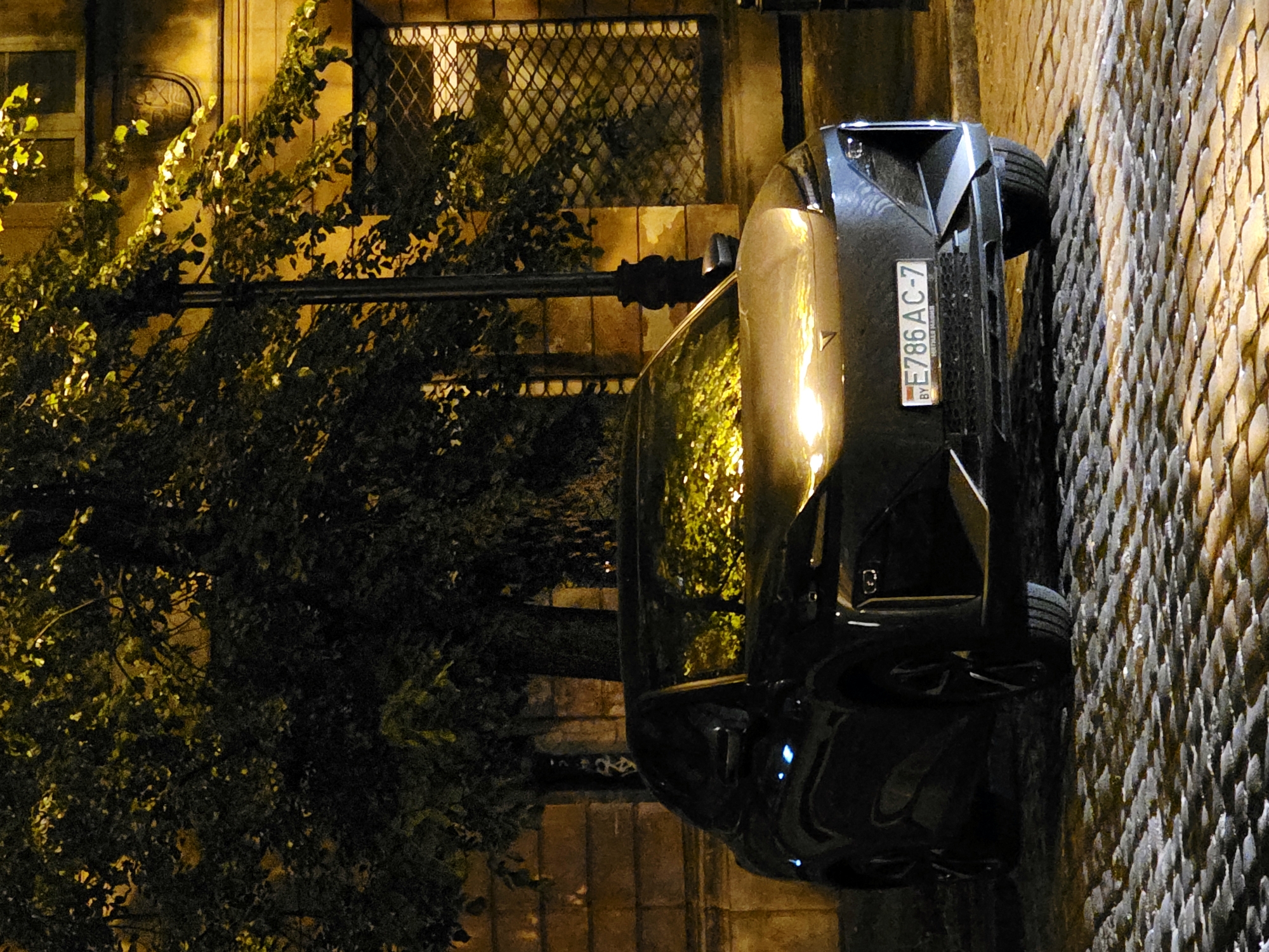 A black car with license plate BY E786 AC-7 is parked on a cobblestone street at night. The car's headlights are on, illuminating the hood and reflecting on the surrounding trees and building. A lamppost stands to the right of the car, and a building with barred windows is visible in the background. The scene appears to be in Warsaw, Poland, given the license plate format which indicates Belarus.