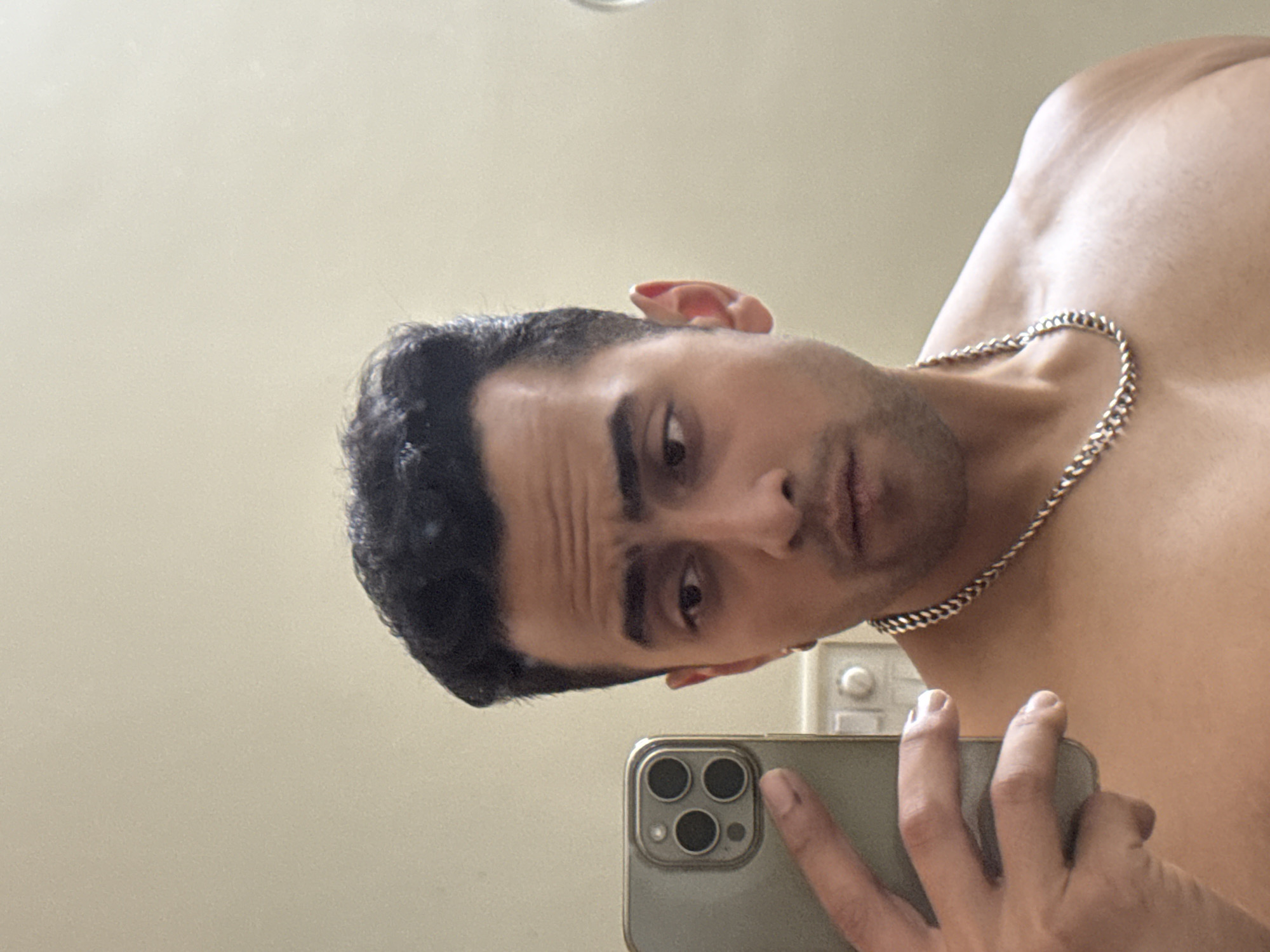 A man takes a selfie in a bathroom with a phone that has three lenses on the back. He is shirtless and wears a silver chain around his neck. He is looking at the camera with a slightly tilted head. The background includes a beige-colored wall and a switch. It is not possible to determine the location or city based on this image alone.
