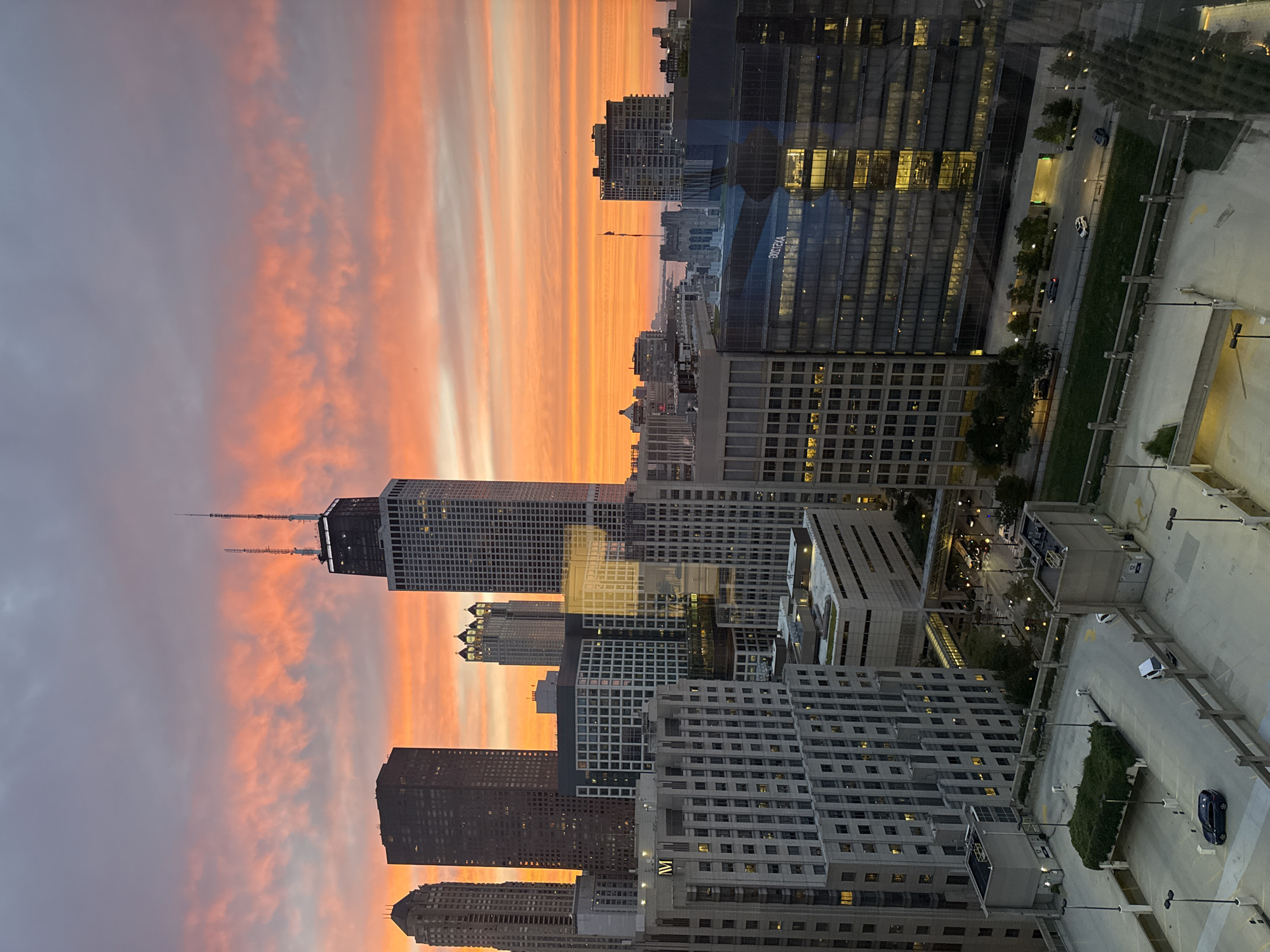 The image captures a sunset over the Chicago skyline. The sky is filled with vibrant orange and pink clouds, contrasting with the silhouetted skyscrapers. Prominent buildings include the John Hancock Center, identifiable by its unique design and antennas, and other modern high-rises. In the foreground, there is a multi-level parking structure with a few cars visible. The overall scene depicts an urban environment during twilight.
