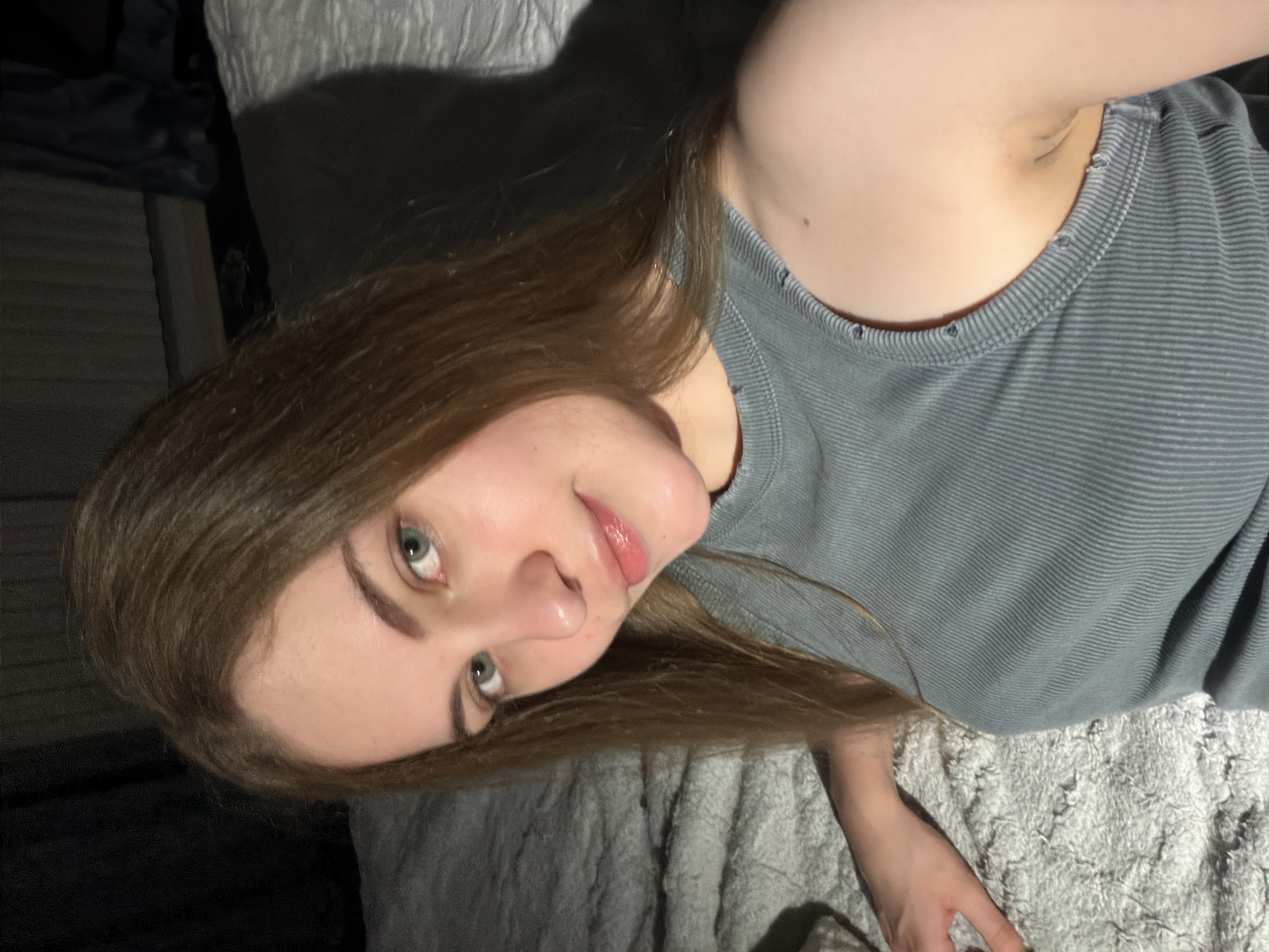 A young woman with long, light brown hair and light blue eyes is shown in a close-up selfie, lying on a textured, off-white blanket. She is wearing a gray ribbed tank top. The lighting in the image is dim, with shadows cast on the right side of her face and body, suggesting an indoor setting, possibly a bedroom. The background is dark and indistinct. There is no indication of the city of Dnipro, Ukraine, or any specific urban location.