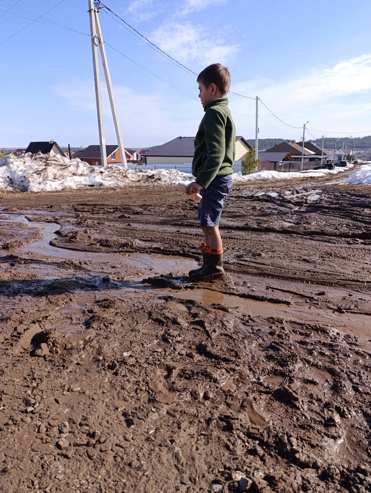A young male child stands in a deeply muddy, unpaved road in a residential area of Chaikovsky, Russia, under a clear, bright blue sky with scattered white clouds. The child, observed in profile facing right, wears a dark green long-sleeved top, dark shorts, and black and orange rain boots. He holds a light-colored, elongated object, likely a stick, in his right hand. The road surface consists of dark brown, wet mud with numerous puddles, extensive tire tracks, and footwear impressions, indicating significant snowmelt. Large, dirty snow piles line the left side of the road, and smaller patches of snow are visible further down on the right. In the background, multiple houses with dark roofs and light-colored facades are visible, along with several concrete utility poles supporting overhead power lines. A dark-colored vehicle is partially visible further along the muddy road. The scene is illuminated by direct sunlight, casting distinct shadows.