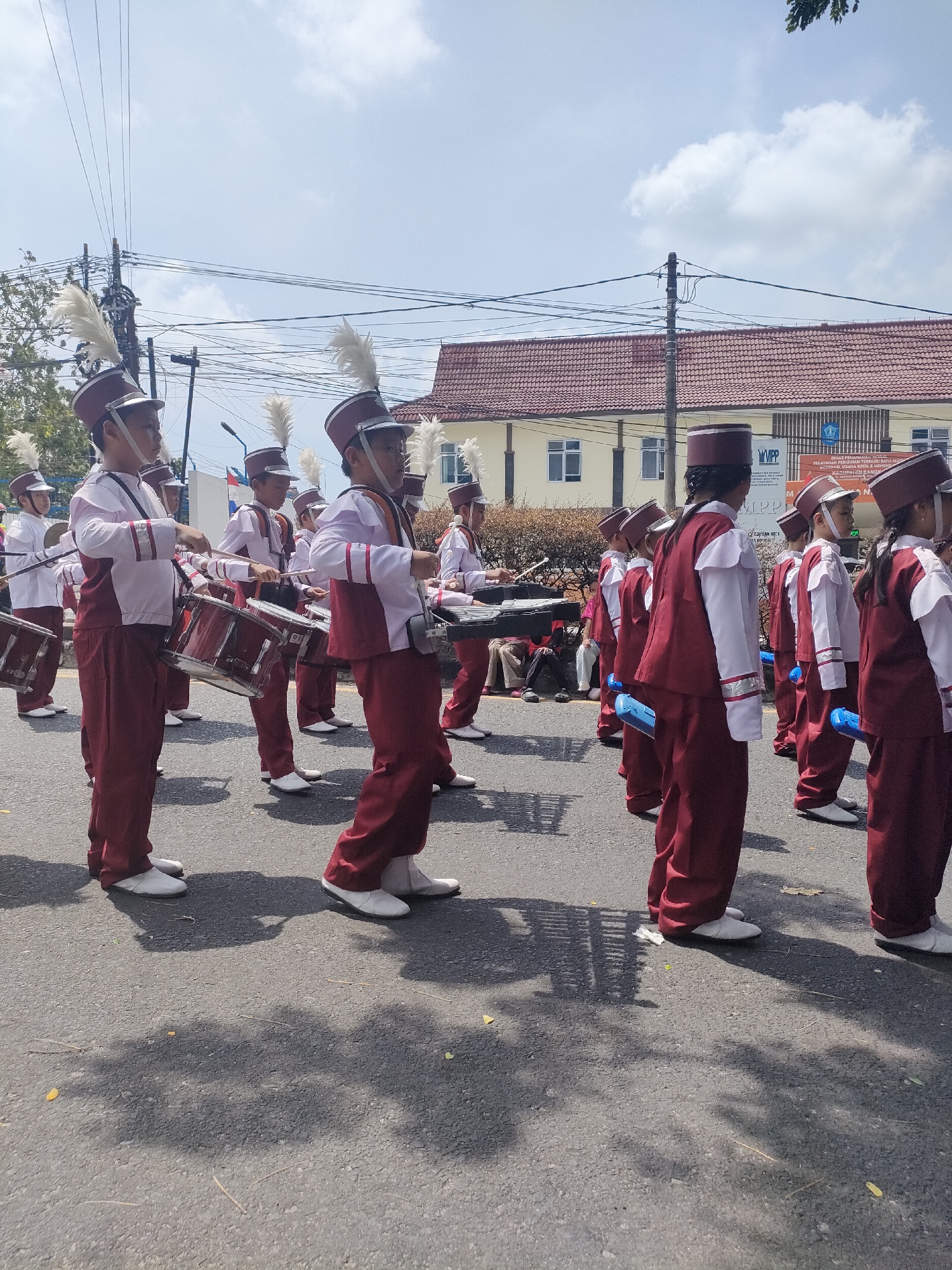 A group of young people in marching band uniforms, predominantly maroon and white, are parading down a street in Sungailiat, Indonesia. The individuals are wearing matching hats with white plumes, white shirts, maroon vests, and maroon pants. Many of them are carrying drums or percussion instruments. The street is paved, and there are trees and utility poles visible on the side of the road. In the background, a building with a red tile roof and several windows can be seen. The sky is blue with scattered clouds. The lighting suggests it is daytime, likely late morning or early afternoon.