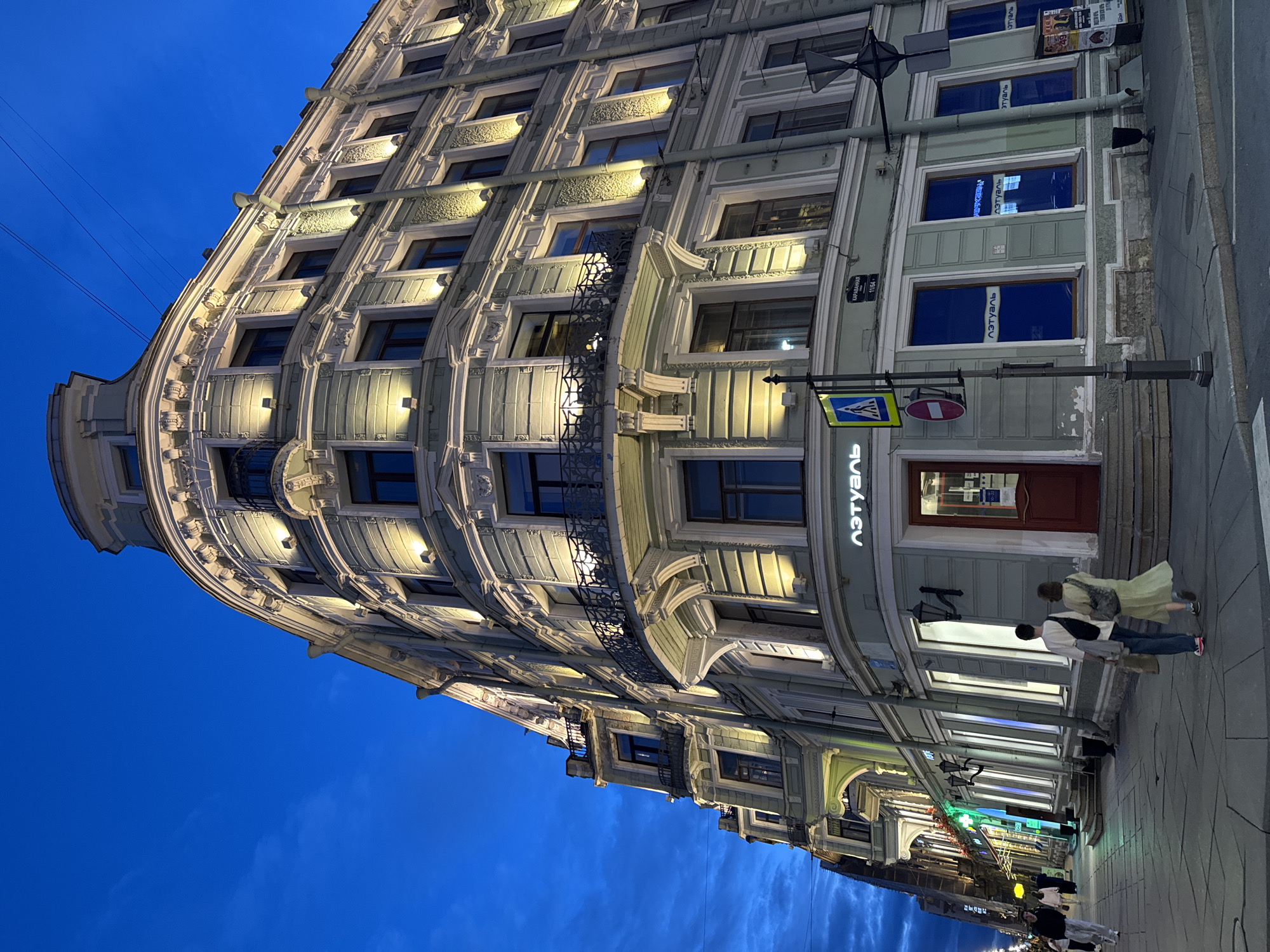The image depicts the exterior of a historic building on a street in Saint Petersburg, Russia, during twilight. The building, illuminated by exterior lights, features ornate architectural details, multiple stories, balconies with decorative railings, and numerous windows. A sign with Russian text, "Лэтуаль," is visible above a storefront. Two individuals, a man and a woman, are walking away from the camera on the sidewalk in front of the building. A street sign displaying pedestrian crossing symbols and a red prohibition sign is mounted on a lamppost. The sky is a deep blue, indicative of the late evening hours, with some clouds present. The sidewalk and street are paved, and other storefronts with illuminated windows are visible further down the street on the left. The overall scene conveys a sense of urban ambiance at dusk.