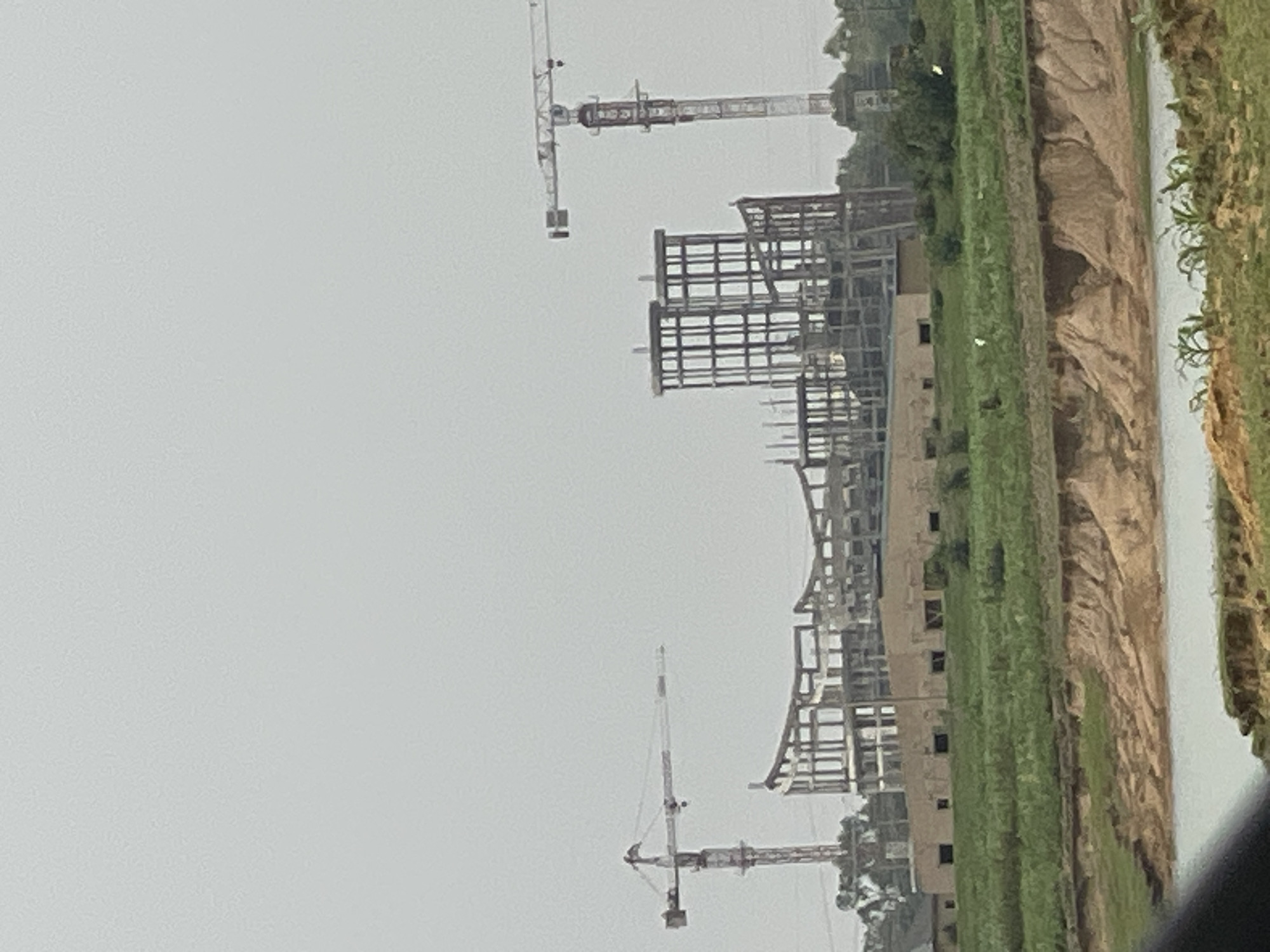 The image depicts a construction site in Nabatum, Nigeria. Several cranes are visible, along with the skeletal structure of a large building under construction. The surrounding environment includes grassy areas and a waterway or large puddle. The sky is overcast, suggesting rainy or humid conditions. No people are directly visible in the scene.