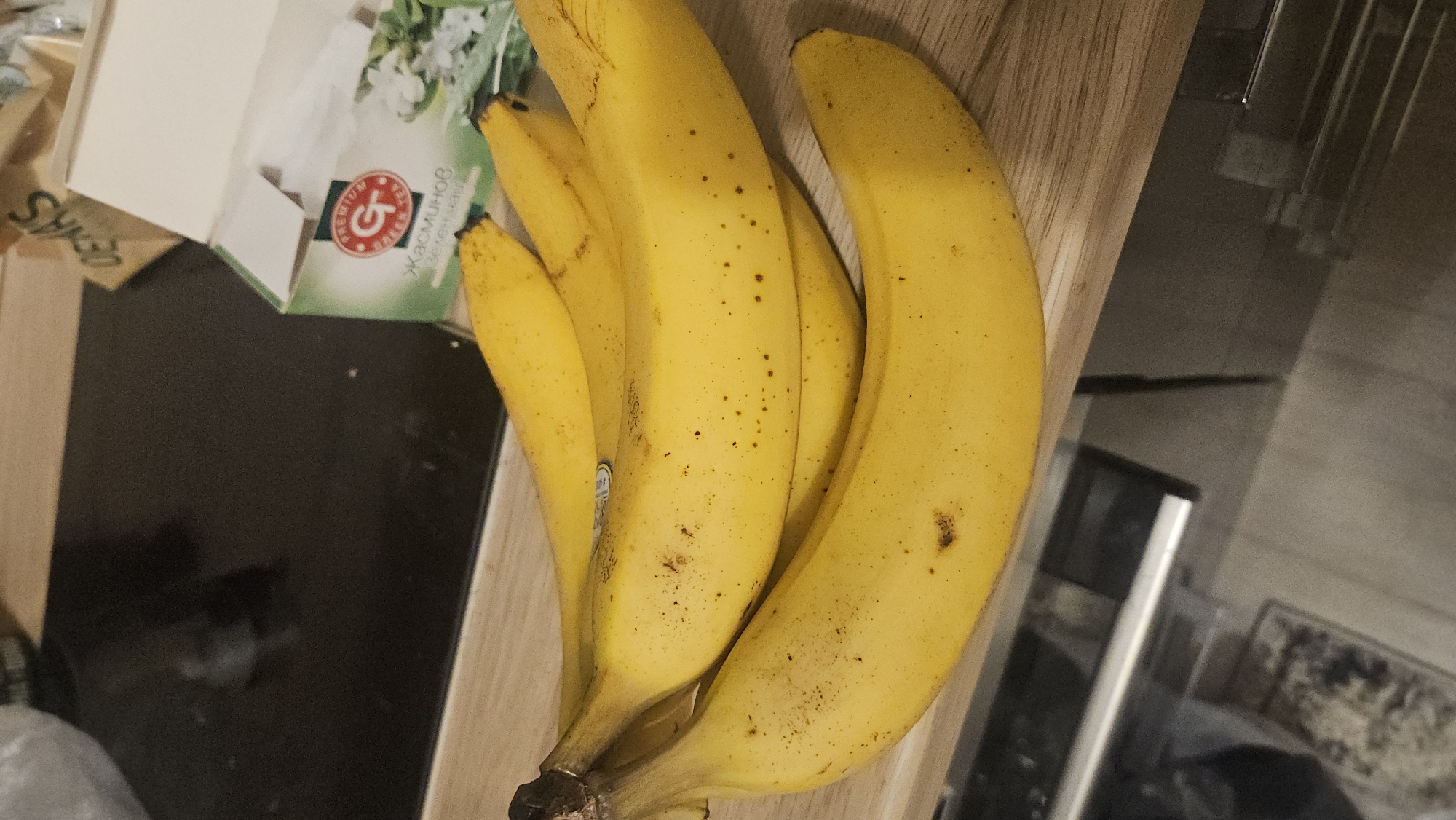 The image shows a bunch of bananas lying on a wooden surface. Next to them is a box with the inscription "ЖАСМИНОВ ЗЕЛЕН ЧАЙ" (Jasmine Green Tea). The background includes parts of a kitchen counter and appliances. The location is impossible to verify as Plovdiv, Bulgaria from the image alone.