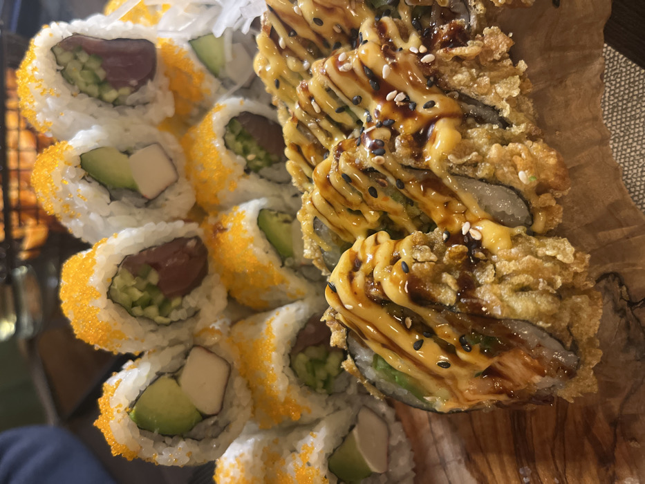 A close-up shot displays two types of sushi rolls arranged on a wooden surface. The left side showcases sushi rolls with orange roe coating, filled with ingredients like tuna, cucumber, avocado, and imitation crab. Adjacent to these are fried sushi rolls drizzled with a creamy yellow sauce and a darker, possibly soy-based, glaze, sprinkled with sesame seeds. The sushi appears freshly prepared and visually appealing.