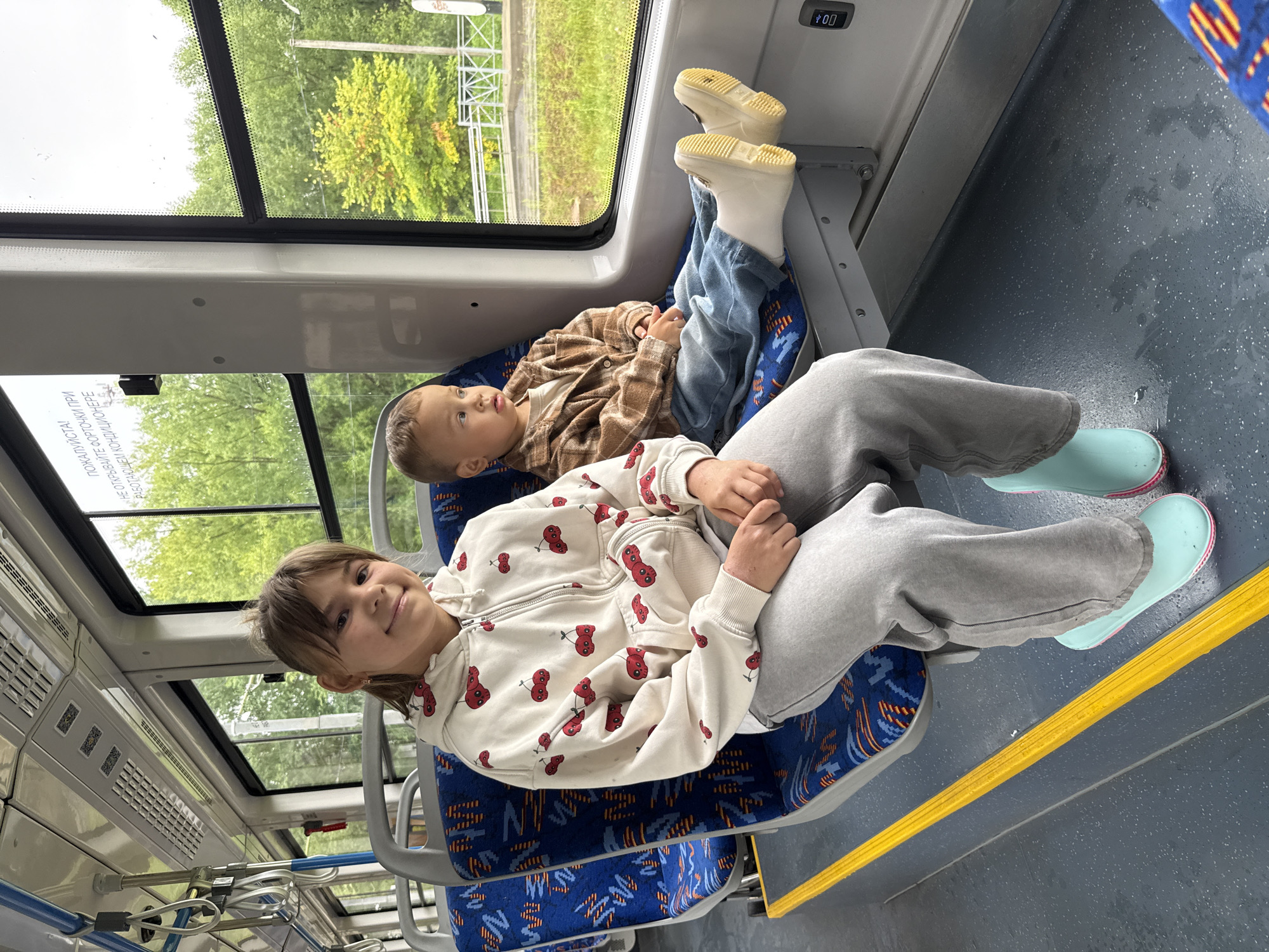 The image captures two children seated on a bus, presumably in Rusanovo, Russia. A young girl with brown hair pulled back in a ponytail is seated on the left, smiling directly at the camera. She wears a white hoodie with a red cherry pattern and gray sweatpants. To her right, a younger boy with light brown hair is looking out the window. He is dressed in a brown plaid shirt and blue jeans, with off-white rubber boots on his feet. The bus interior features blue patterned seats and a yellow stripe on the floor. Through the windows, green trees and foliage are visible, suggesting an outdoor, possibly rural or suburban, setting. The lighting indicates it is daytime, with an overcast sky.