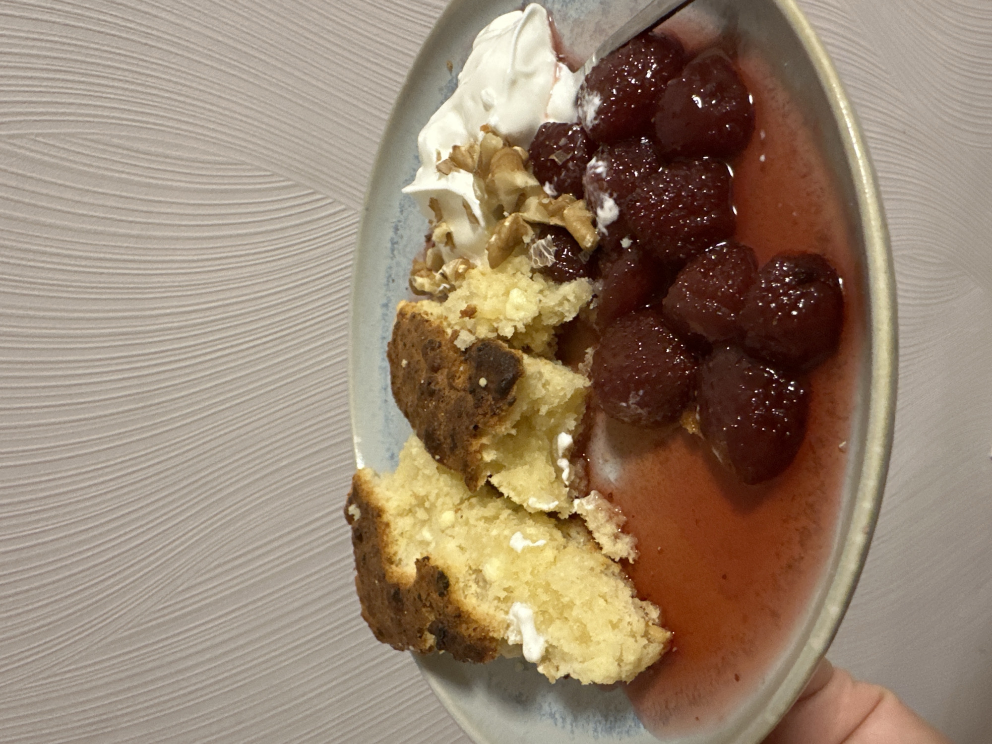 A plate of food is shown in the foreground, with a textured, light-colored wall in the background. The plate contains pieces of a golden-brown cake, some of which appear to be slightly overcooked. On the right side of the plate, there are cooked strawberries in a red syrup, a dollop of white cream, and chopped walnuts. A hand is visible on the bottom left, holding the plate. The scene appears to be indoors, likely in a home setting in Khimki, Russia.
