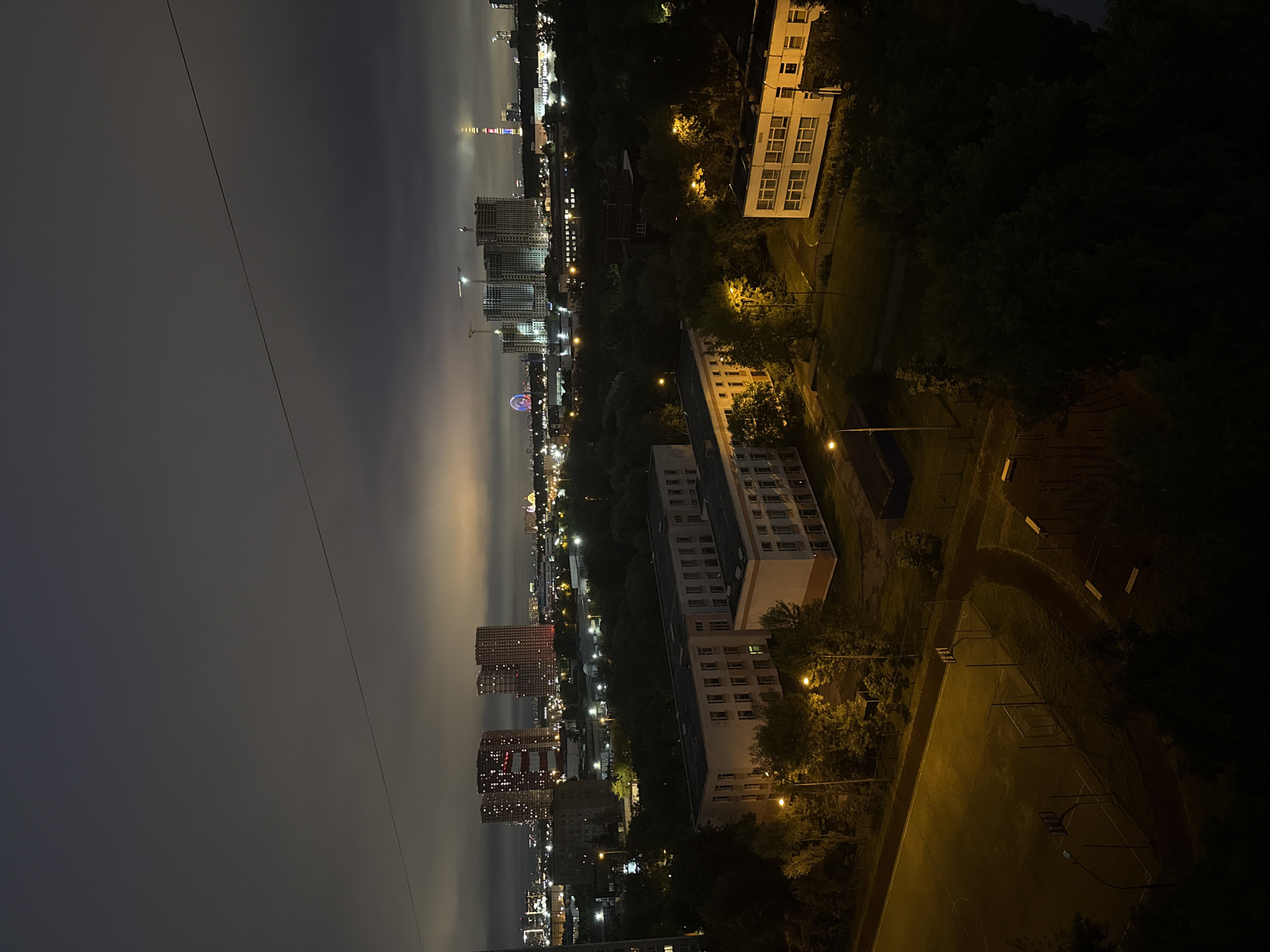 This is a nighttime aerial photograph taken from an elevated vantage point, looking out over a suburban area of Moscow, Russia. The sky is dark and overcast, with a faint glow on the horizon suggesting city lights and possibly some pollution or cloud illumination. A single power line stretches diagonally across the upper portion of the frame.
In the foreground, a sports field, likely a basketball court or multi-purpose area, is illuminated by artificial lighting. The field is bordered by trees and a low fence. Adjacent to the sports field are two school-like buildings with multiple windows, illuminated from within. These buildings appear to be low-rise, with flat roofs.
Further into the middle ground, a dense line of trees separates the immediate buildings from the more distant urban landscape. Beyond the trees, a variety of buildings are visible, including residential towers with illuminated windows, some appearing to be under construction with visible cranes. A Ferris wheel, lit with colorful lights, is a prominent feature in the mid-distance. A tall, illuminated tower, possibly a broadcast tower, stands out in the far distance, distinguished by its red and white colored bands. The overall impression is of a developed urban area at night, with a mix of residential, recreational, and construction activity. There are no visible people in the frame.