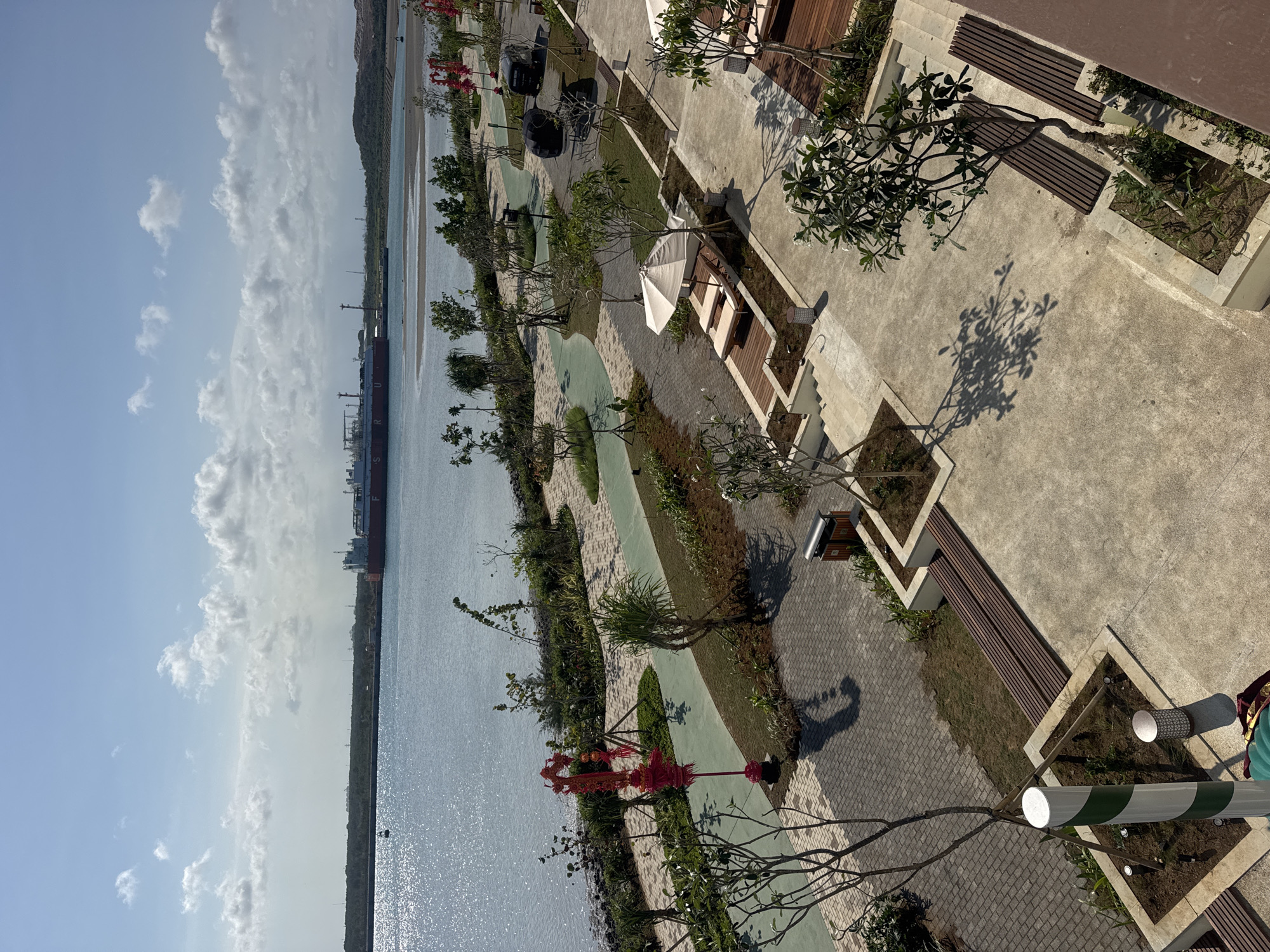 The image shows an outdoor area with a body of water on the left and a paved walkway with benches and lounge chairs on the right. In the water, a large red and white ship is visible in the distance, along with a smaller boat. The walkway area features several trees, some with lounge chairs and umbrellas set up. There are also two black vehicles parked on the right side of the walkway. The sky is blue with scattered white clouds. The location is identified as Unknown, Indonesia.