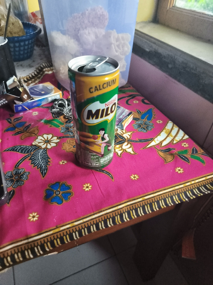 A can of Milo chocolate drink sits on a pink tablecloth with a floral pattern. The can is green and yellow and has a picture of a woman playing basketball. The table is made of wood and has a black and white border around the edge. The table is set in a room with a pink wall and a window with a view of green trees. The coordinates provided are for the city of Majalengka, Indonesia. There is a large plastic container containing a purple substance that appears to be ice cream. The container is behind the can of Milo. Several items are partially visible on the table, including a plastic bag, cigarette lighter, and other various objects. There is a brown, empty package, partially visible at the left edge of the table. There is also a small, dark blue, round object with a white label.
