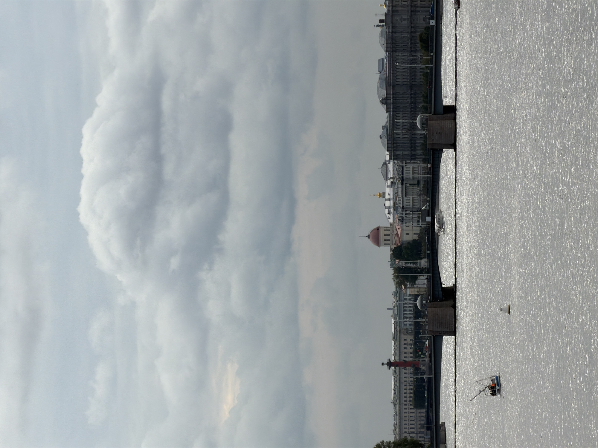 The image captures a view of the Neva River in Saint Petersburg, Russia, with a bridge spanning across it. The sky is overcast with dramatic, heavy clouds. In the foreground, the shimmering surface of the Neva River is visible, with a small boat carrying several people sailing on it. A white buoy floats in the water. In the background, the St. Petersburg skyline is depicted, featuring various historical buildings, including a prominent red columned building, a domed structure with a red roof, and long, classical facades. The bridge itself is a substantial structure with stone pillars and a dark roadway. The overall atmosphere is somber due to the cloudy weather, yet the scene is vibrant with the movement of the boat and the reflections on the water.