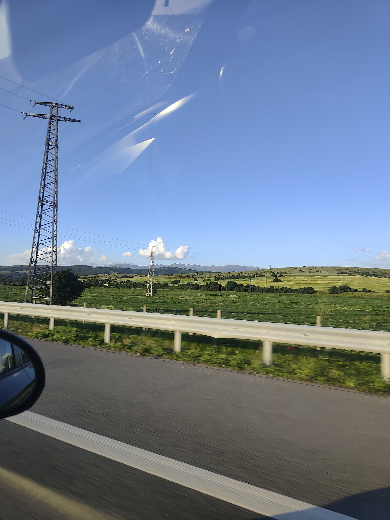 The image depicts a view from inside a moving vehicle, likely on a highway near Dolna Dikanya, Bulgaria. The sky is clear blue with some light cloud cover. The landscape features rolling green hills with patches of trees and cultivated fields. Two electrical transmission towers are visible in the distance. A white guardrail runs along the side of the road. A portion of the car's side mirror is visible in the lower left corner. There are smudges or reflections on the windshield of the car.
