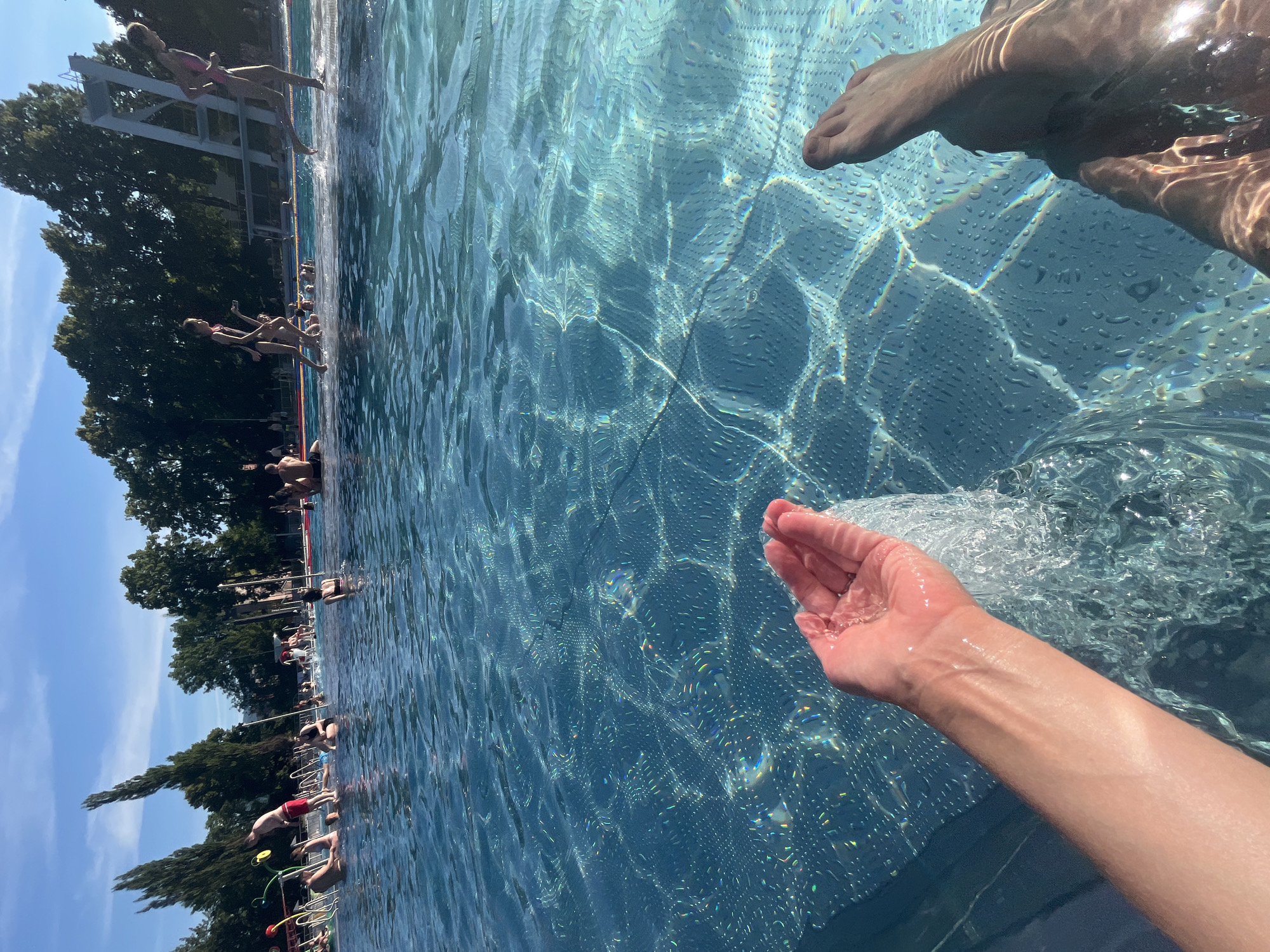 A first-person perspective shot of a person's arm and hand holding water in a swimming pool. The hand is positioned to let the water flow back into the pool, creating a splash. In the background, several people are visible in the pool, some swimming and others relaxing. The pool water is clear and blue, with sunlight reflecting off its surface, creating shimmering patterns on the tiled bottom. Trees and a diving platform are visible in the distance, under a clear blue sky.