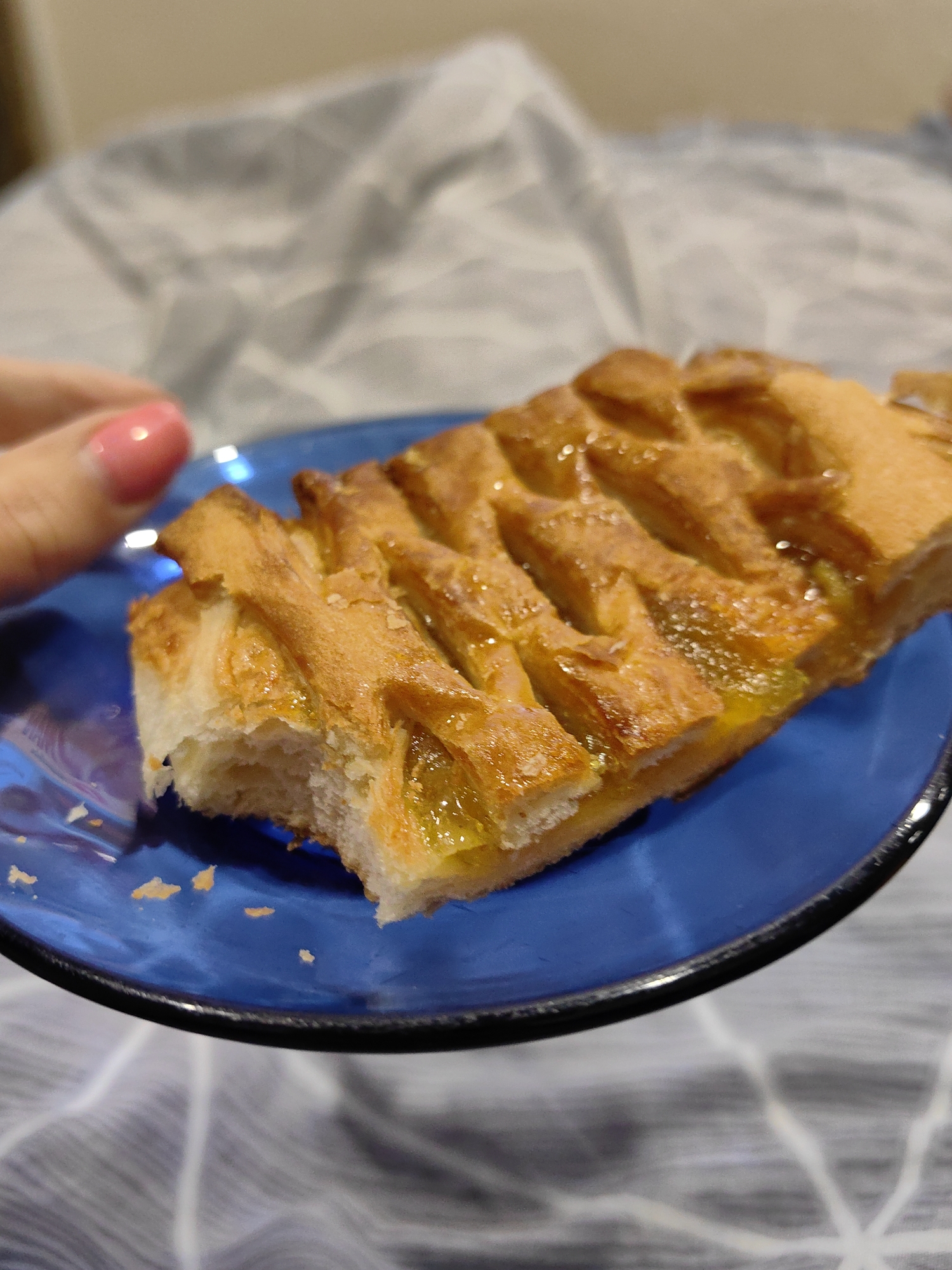 A slice of pie with a lattice top, filled with what appears to be apricot jam, sits on a blue plate. A person's hand with a pink fingernail is visible to the left, reaching towards the pie. The setting is a blurred background, suggesting an indoor environment. The image does not provide information about its location in Fanipal, Belarus.