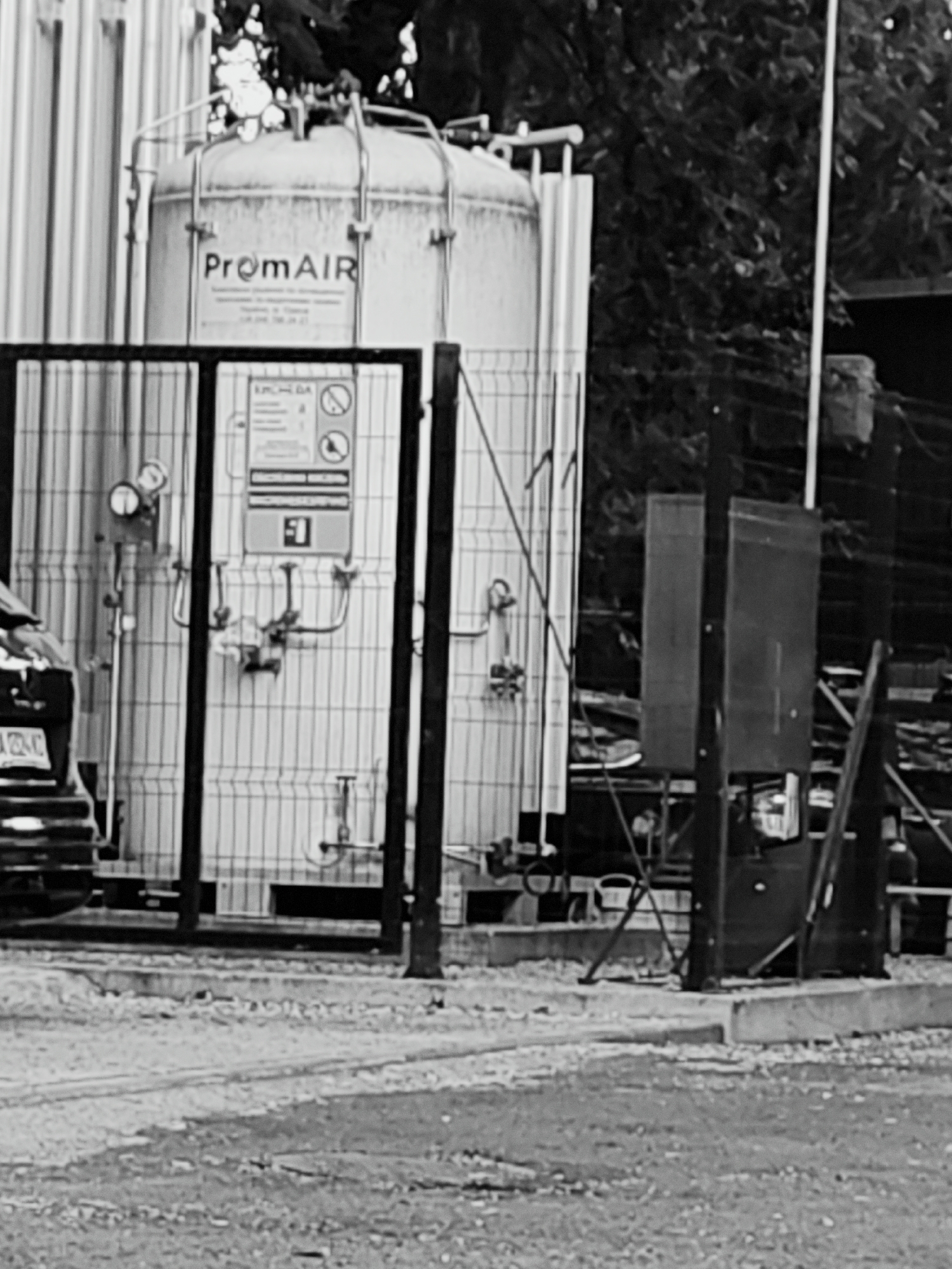 The image shows an industrial scene in Odesa, Ukraine. In the foreground, a chain-link fence encloses a large, vertical cylindrical tank. The tank has the "PromAIR" logo visible on its side. Attached to the tank are various pipes, valves, and gauges. There is also a safety sign with symbols and text. To the left of the tank, part of a dark-colored vehicle is visible. Behind the fence and tank, the scene is blurred, showing trees and other industrial structures. The ground in the foreground is rough and appears to be a dirt or gravel surface. The overall impression is that of an industrial facility likely involved in gas storage or processing.