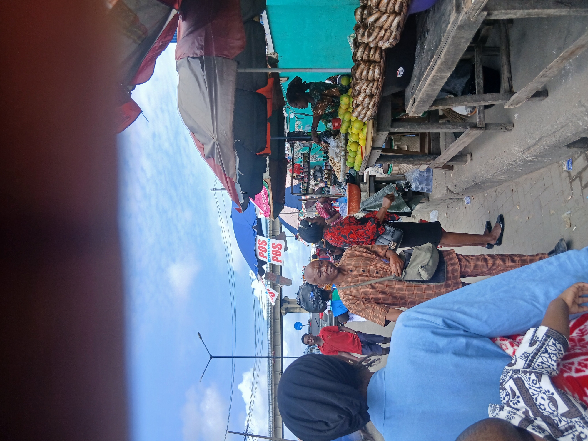 The image depicts a lively outdoor market scene in Aganí, Nigeria. Several people are visible, dressed in various casual and traditional attire. In the foreground, a person wearing a blue outfit is partially visible, sitting. To their right, another person in a patterned white and black garment is also seated. In the middle ground, a man in a brown and orange checkered shirt and pants is looking towards the camera. He is holding a stick or a pole. A woman in a red and black patterned top is standing behind him. Further back, a woman in a green patterned dress is attending to a stall filled with produce, including many green mangoes and what appear to be dried fish hanging above. The market is set up under awnings and makeshift shelters, suggesting it's an active marketplace.
Objects in the scene include market stalls, produce (mangoes, possibly other fruits), dried fish, bags, umbrellas (one blue with "POS POS" visible text), and a street light. The ground appears to be paved or a dirt path. In the background, there's a glimpse of a bridge or overhead structure and a street with some traffic.
The time of day appears to be daytime, with bright natural light. The sky is visible and mostly clear with some clouds. The overall atmosphere seems busy and active, typical of a bustling market. The people's expressions are varied, with some looking directly at the camera. There are no overt signs of distress or joy, but the general feel is one of everyday commerce. The text "POS POS" is clearly visible on a blue umbrella, likely indicating Point of Sale services are available.