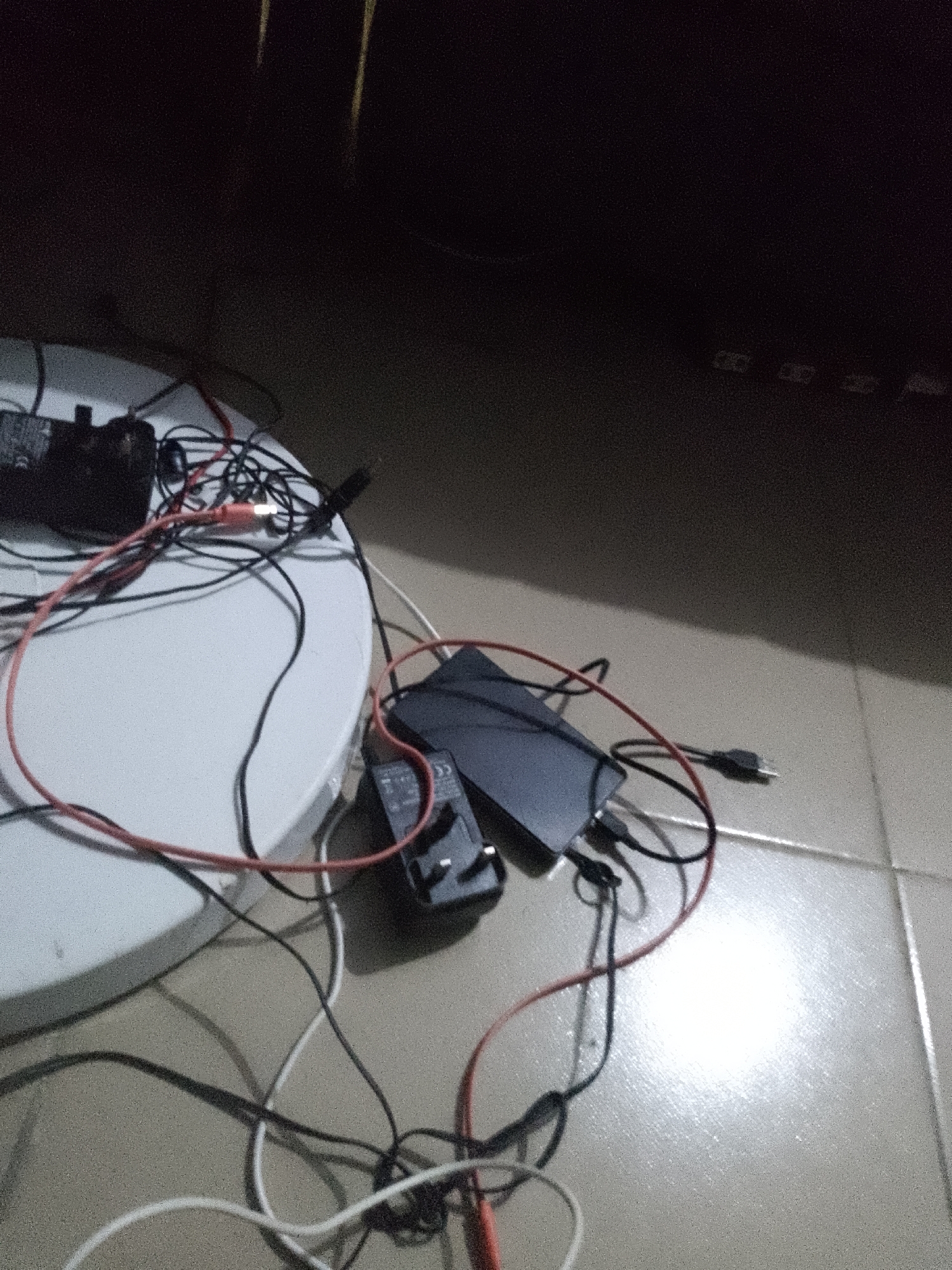 The image depicts a cluttered arrangement of electronic devices and cables on a white circular surface, possibly a table, and the tiled floor in what appears to be a dimly lit interior. Several black power adapters are visible, one connected to a black rectangular device, likely a power bank or external hard drive. Red and black wires snake across the scene, some connecting the adapters to the devices, while white cables also contribute to the disarray. The overall impression is one of disorganization, with multiple charging or connection points seemingly in use or set up. The setting is not identifiable as being in the city of Jauro Yinu, Nigeria; the image provides no such contextual information.