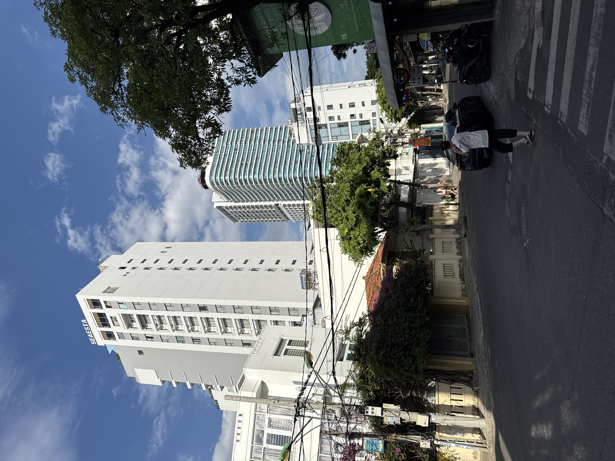 This image depicts a street scene in Nha Trang, Vietnam, under a bright, cloudy sky. In the background, two tall buildings dominate the skyline: a white, modern high-rise labeled "SEAEASTA" with a contemporary design, and another building with a distinctive curved facade featuring numerous horizontal green-tinted windows. In the foreground, a paved road with a pedestrian crossing is visible. A person wearing a white t-shirt and dark pants is standing on the right side of the road, facing away from the camera. Several parked motorcycles are also present. The street is lined with trees and smaller buildings, including one with a tiled roof and a yellow facade. Electrical wires crisscross the scene, suggesting an urban environment with well-established infrastructure. The overall atmosphere is one of a typical day in a bustling Vietnamese city, with a blend of modern architecture and older structures.