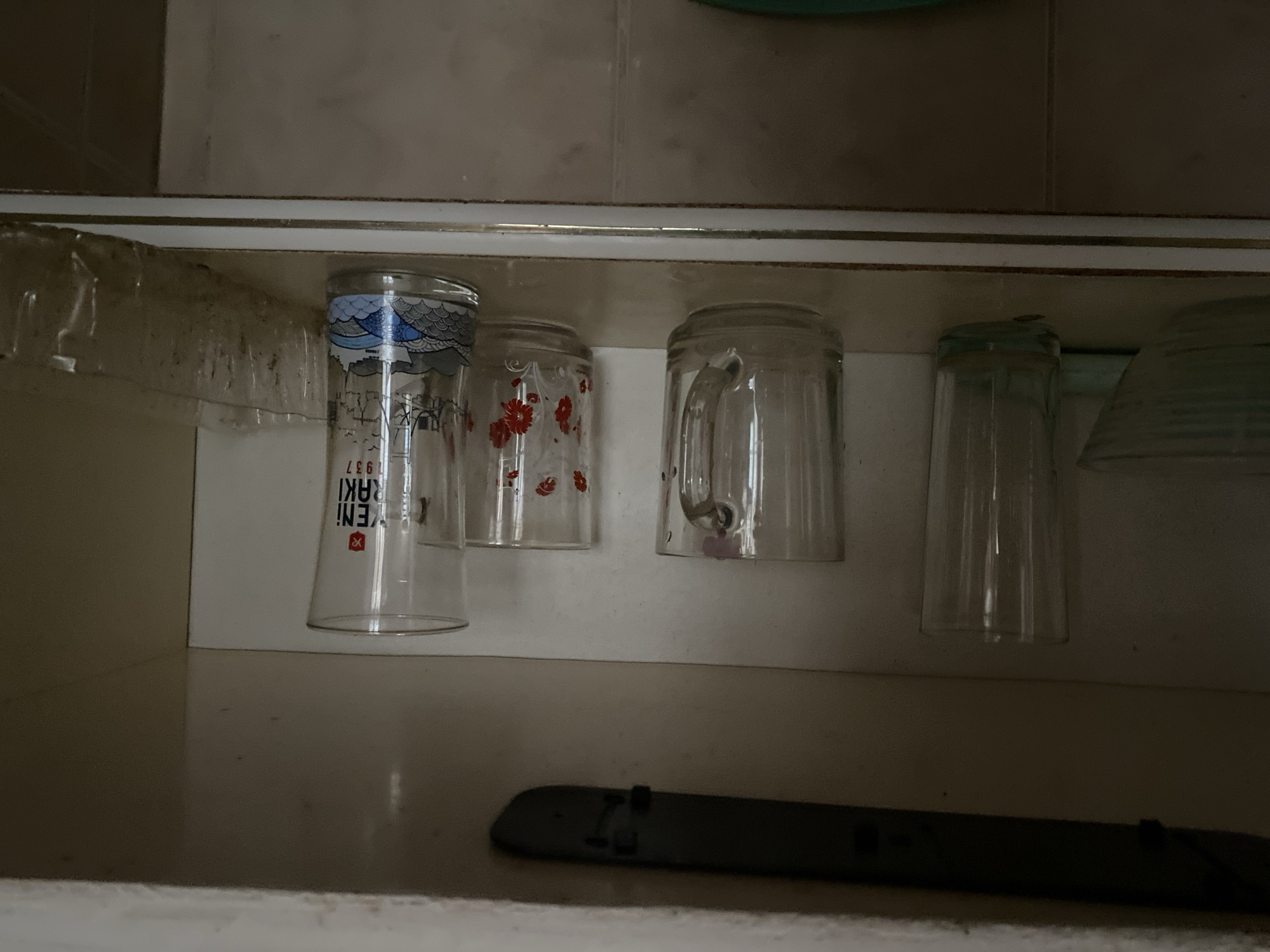 A wide-angle, eye-level shot captures the interior of a dimly lit kitchen cabinet in Sokhumi, Abkhazia. The cabinet shelf is lined with various glassware, including a tall, clear glass on the far left, a mug with a handle, a smaller glass with red floral patterns, and a taller glass adorned with a depiction of a boat on waves and text that reads "YENI RAKI 1937". To the right of these glasses is a dirty, transparent plastic container. The background wall of the cabinet is off-white, and the shelf itself has a white trim. Above the shelf, a dark, horizontal object, possibly a light fixture or a cabinet support, is visible. The scene is consistent with household storage, with the objects appearing clean but showing signs of normal use or being somewhat dusty.