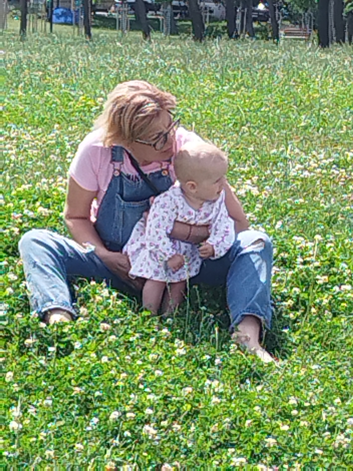 The image depicts a woman and a baby sitting on a grassy area with small white flowers. The woman is wearing a pink t-shirt and denim overalls, with blonde hair and glasses. She is holding the baby, who is wearing a light-colored dress with a floral pattern. They are in a park-like setting with trees in the background. The scene appears to be during the daytime, with sunlight illuminating the grass and flowers. This image was taken in "La Plaja" in Constanța, Romania, specifically in the park adjacent to the beach.