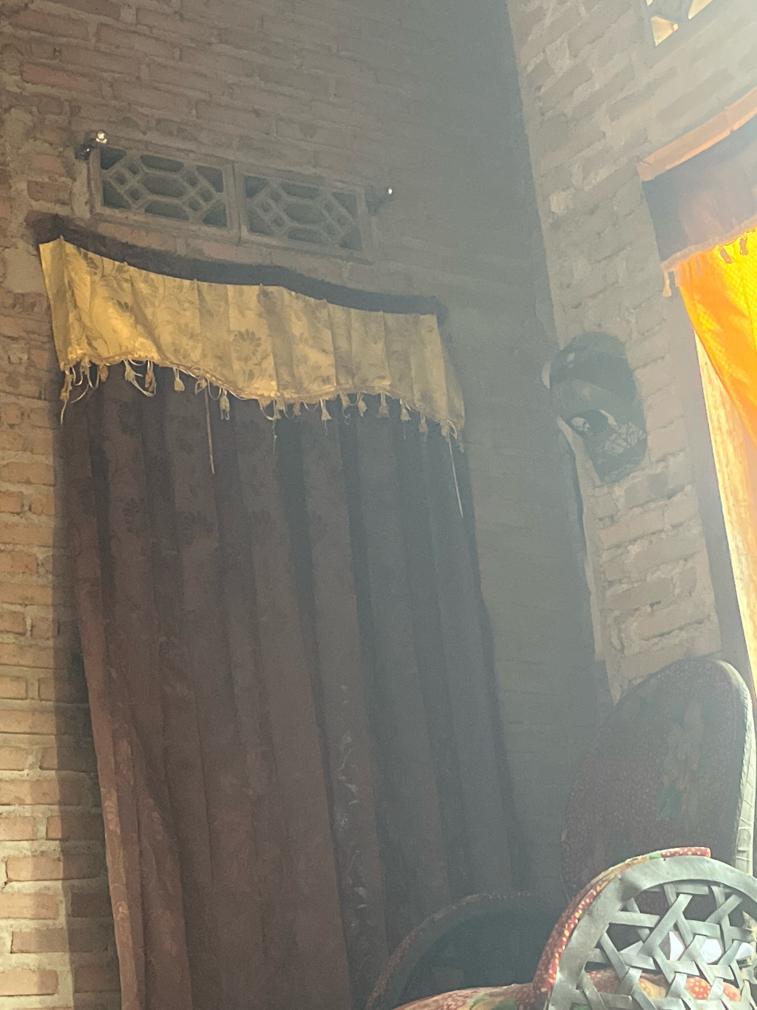 This image depicts an interior scene, likely a room, with brick walls. A dark brown curtain hangs prominently, partially covering a window area which features decorative concrete blocks. Above the curtain, a yellow valance with a floral pattern and fringe is visible. To the right, part of an orange curtain or fabric is illuminated by sunlight. In the background, a dark object resembling a decorative mask is affixed to the brick wall. In the foreground, a portion of a chair with a patterned upholstery and a woven seat is visible. The overall lighting suggests a daytime setting with sunlight streaming in from the right. The image does not provide specific information about the city of Sidomulyo, Indonesia, beyond its potential location.