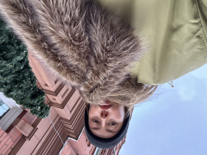 A low-angle photograph captures a young adult female looking directly into the camera in London, United Kingdom, on an overcast day. She wears a black knitted beanie and an olive green jacket featuring a prominent brown faux-fur collar or stole. Her brown hair is visible beneath the beanie. The background features a multi-story red brick building with distinctive architectural cornices and ledges. To the subject's right, a vertical evergreen decoration, adorned with small white lights, is affixed to the building, suggesting a festive or winter period. The upper left portion of the frame shows an expanse of cloudy sky.