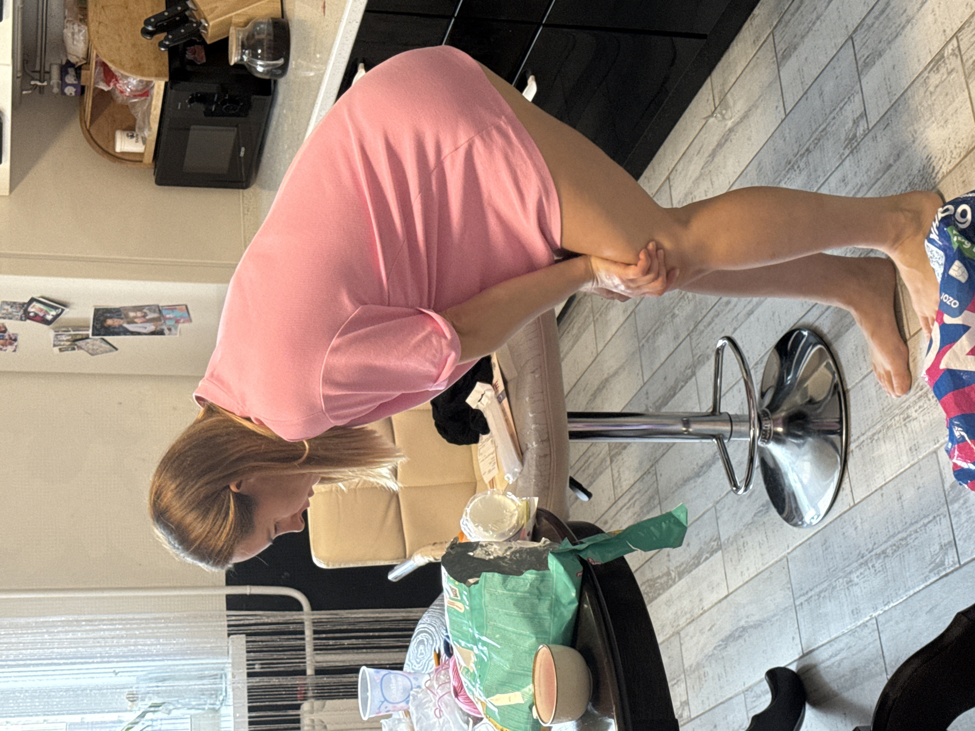A fair-skinned woman in a pink dress bends forward from a barstool. Her hands are clasped together in front of her legs. She has light brown hair. The barstool is upholstered in a light color, and has a chrome base. On a table in front of her is a trash bag, disposable cups, and other discarded items. The flooring appears to be light gray tile. There is a window in the background with vertical blinds. Black cabinets are visible in the background. The location is an interior. The city is not visible in the image.