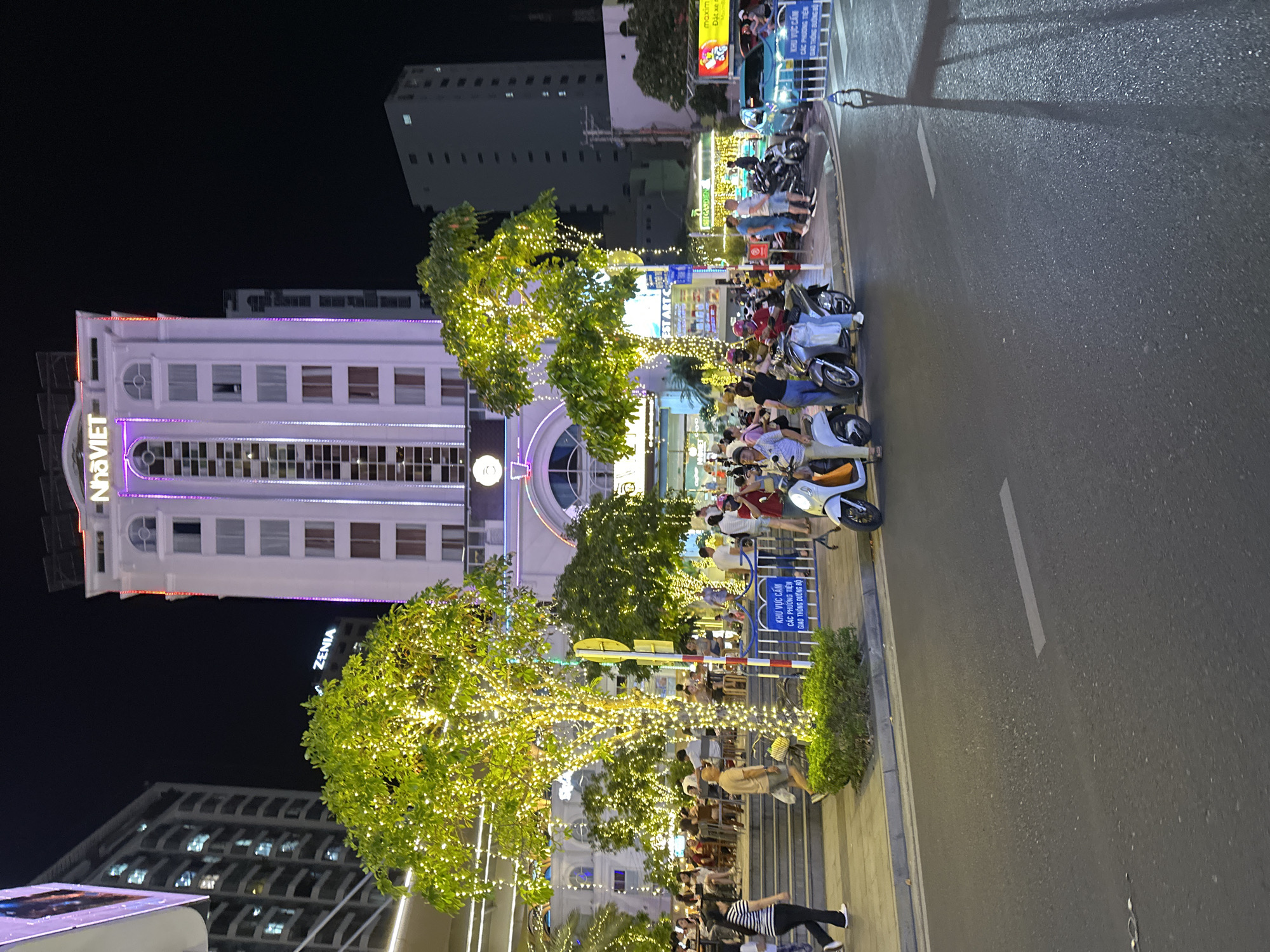 This is a night-time street scene in Nha Trang, Vietnam, likely near the city center given the presence of a large, illuminated building and a bustling pedestrian area. The focal point is a tall, white building with "NhaVIET" written in prominent, illuminated letters at its apex. The building is adorned with purple and red lights, suggesting it's a hotel or commercial establishment. In front of it, a tree is heavily decorated with numerous small, warm-toned lights, creating a festive atmosphere.
The street in the foreground is paved and appears to be a road with markings. To the left, there's a sidewalk with a group of people walking and sitting at outdoor tables, indicating a commercial or dining area. Several motorcycles and scooters are parked and in motion on the street and sidewalk, typical of urban Vietnamese transportation. A blue sedan is also visible.
In the midground, there's a white barrier with text in Vietnamese, and a sign with a yellow circular element. The presence of multiple people, outdoor seating, and illuminated trees suggests a vibrant urban environment, possibly a promenade or a lively commercial district. The overall impression is that of a busy evening in a Vietnamese city, with elements of both modern architecture and pedestrian-friendly street life.
