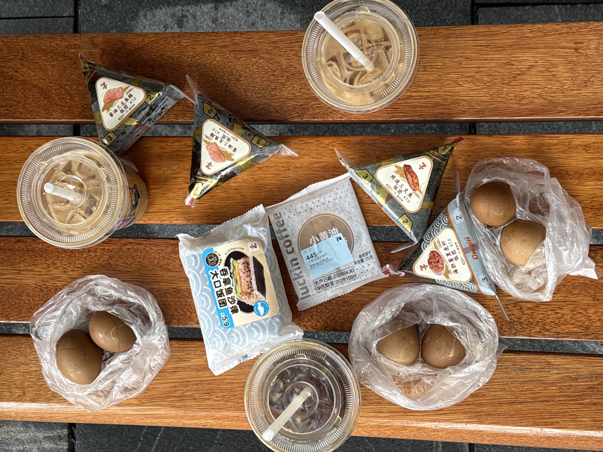 The image shows a collection of food and drinks spread across a wooden bench, likely outdoors.
There are several onigiri (Japanese rice balls) in their packaging, including some with tuna salad filling and others with salmon. Two small packages of cookies from Luckin Coffee, labeled "Small Butter Cookies" are also visible. Three plastic bags each contain two brown-colored boiled eggs. There are also three cups of iced coffee or similar beverages, each with a straw.
The scene is set on a bench made of parallel wooden slats, and the shadow on the bottom implies an outdoor location. The presence of Chinese packaging and labels, including the mention of "Luckin Coffee" and food prices in Chinese currency, suggests that the location is in China. The OCR results also mention Tianhe District, China.