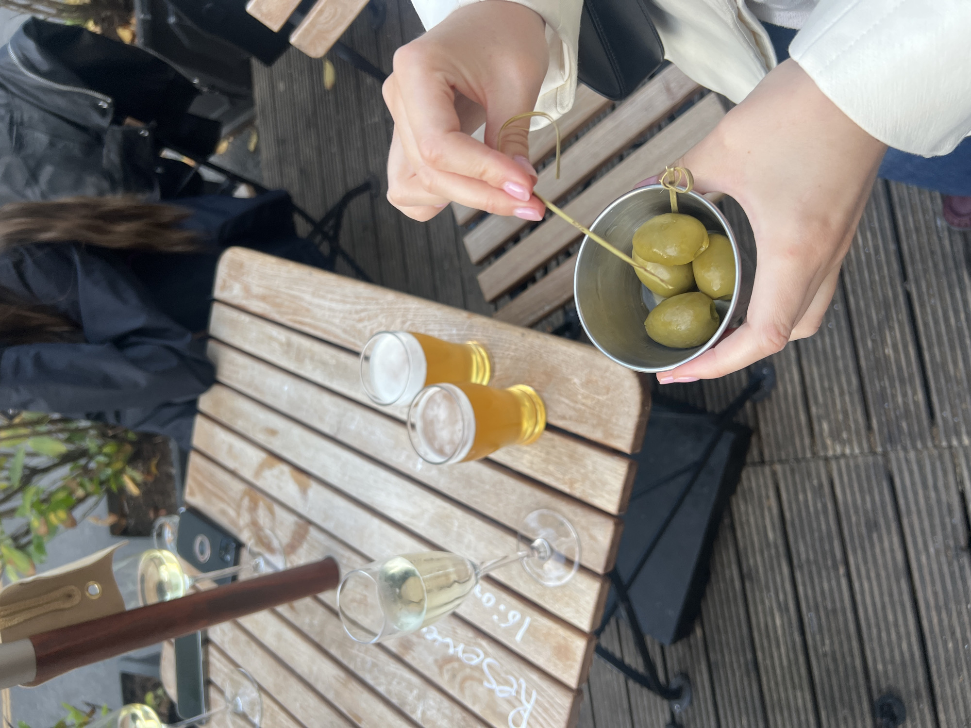 The image shows a view from above of a table in what appears to be an outdoor eating area. Two beers and two glasses of sparkling wine are visible. A hand holds a small metal cup of olives with a toothpick. The word "Reserved" is partially visible on the table. The setting appears to be a wooden deck or patio. Without additional visual clues, it is impossible to confirm the location as London or anywhere else in the United Kingdom.
