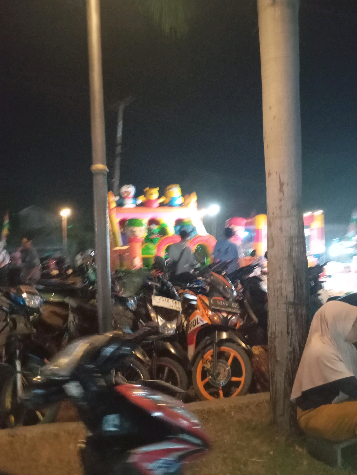 The image depicts a nighttime scene in Kecamatan Haurgeulis, Indonesia, likely at a public gathering or festival. In the foreground, several motorcycles are parked, with one in sharp focus showcasing an orange and black color scheme and an orange rimmed wheel. The license plate of this motorcycle is partially visible, reading "E 5969 OZT". To the right, a person wearing a light-colored hijab and yellow pants is seated. In the background, a large inflatable attraction, possibly a bouncy castle, is illuminated and features cartoon characters on top, including what appear to be Minions and a character resembling Doraemon. Other individuals are visible around the inflatable, some appearing to be children or attendees. A tall, light-colored pole, possibly a lamppost, stands prominently in the left-center of the frame, and a large tree trunk is on the right. The overall atmosphere suggests a lively evening event.