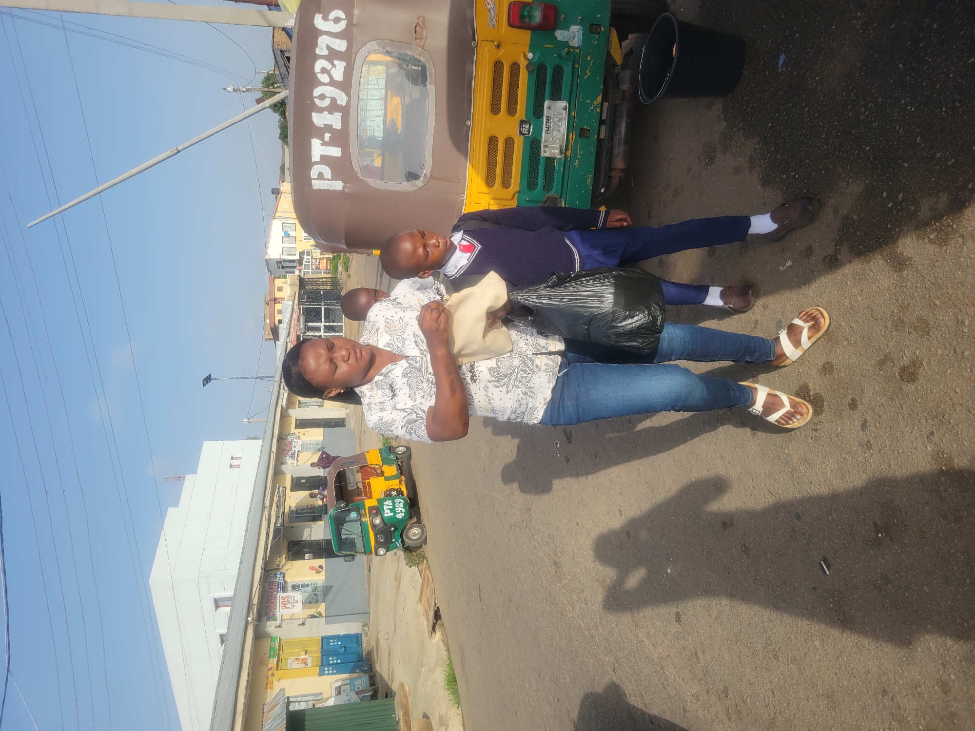 The image depicts a street scene in Jos, Nigeria, with a woman and a young boy standing next to a parked three-wheeled vehicle, locally known as a "Keke Napep." The woman is wearing a light-colored patterned shirt and jeans, holding two bags (one black, one light-colored). The boy is wearing a school uniform, consisting of a dark blue sweater over a white shirt and dark blue trousers. The Keke Napep is painted in brown, green, and yellow, with the registration number PT-19276 and PLATEAU state visible on its plates. A second green and yellow Keke Napep is visible further down the street. The background shows buildings and power lines under a blue sky. The road is paved with asphalt.