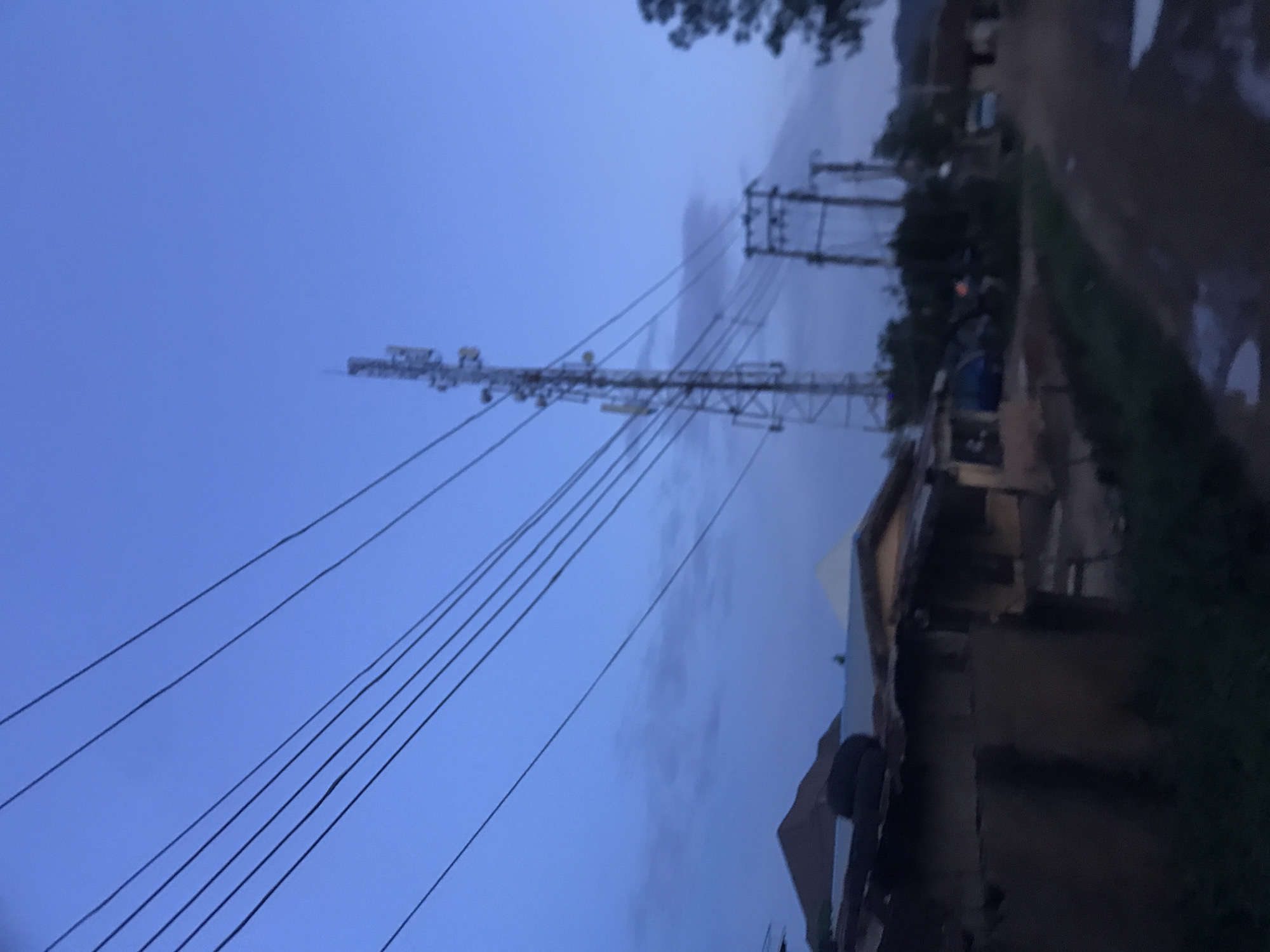 The image portrays an outdoor scene in what is identified as Rafin Par, Nigeria, observed during twilight, characterized by a deep blue sky and diffuse, low-level illumination.
The upper two-thirds of the frame features a clear, deep blue sky with sparse, horizontally elongated grey clouds visible in the mid-left area. Several dark, parallel utility lines traverse diagonally from the upper left, converging towards a tall, metallic lattice-style telecommunications tower positioned centrally. This tower is equipped with multiple antennae and dishes at various heights. To the right of the main tower, a more robust utility pole or smaller transmission tower with cross arms and insulators supports additional power lines, with another similar structure partially visible further to the right. A dark, indistinct tree mass partially occupies the far right edge of the frame.
The lower third of the image depicts ground-level structures and a dirt road. On the left foreground, several low-rise buildings are visible. The most prominent is a dark, earthen or mud-walled structure with a dark, possibly corrugated metal roof, upon which two dark, circular objects (likely tires) are resting. Behind this, a building with lighter-colored walls and a gabled roof is partially seen. In the distant left background, a distinct brown, conical hill or mountain shape rises against the sky, with another light-colored, angular roof structure visible further right.
An unpaved road extends from the lower central foreground towards the right background. Several bright reflections on the road surface indicate the presence of puddles. Dark, blurry vegetation lines the edges of the road. In the far distance along the road, indistinct structures and the faint outline of a white vehicle are discernible on the right. No individuals are visible, and no specific actions or interactions are depicted.