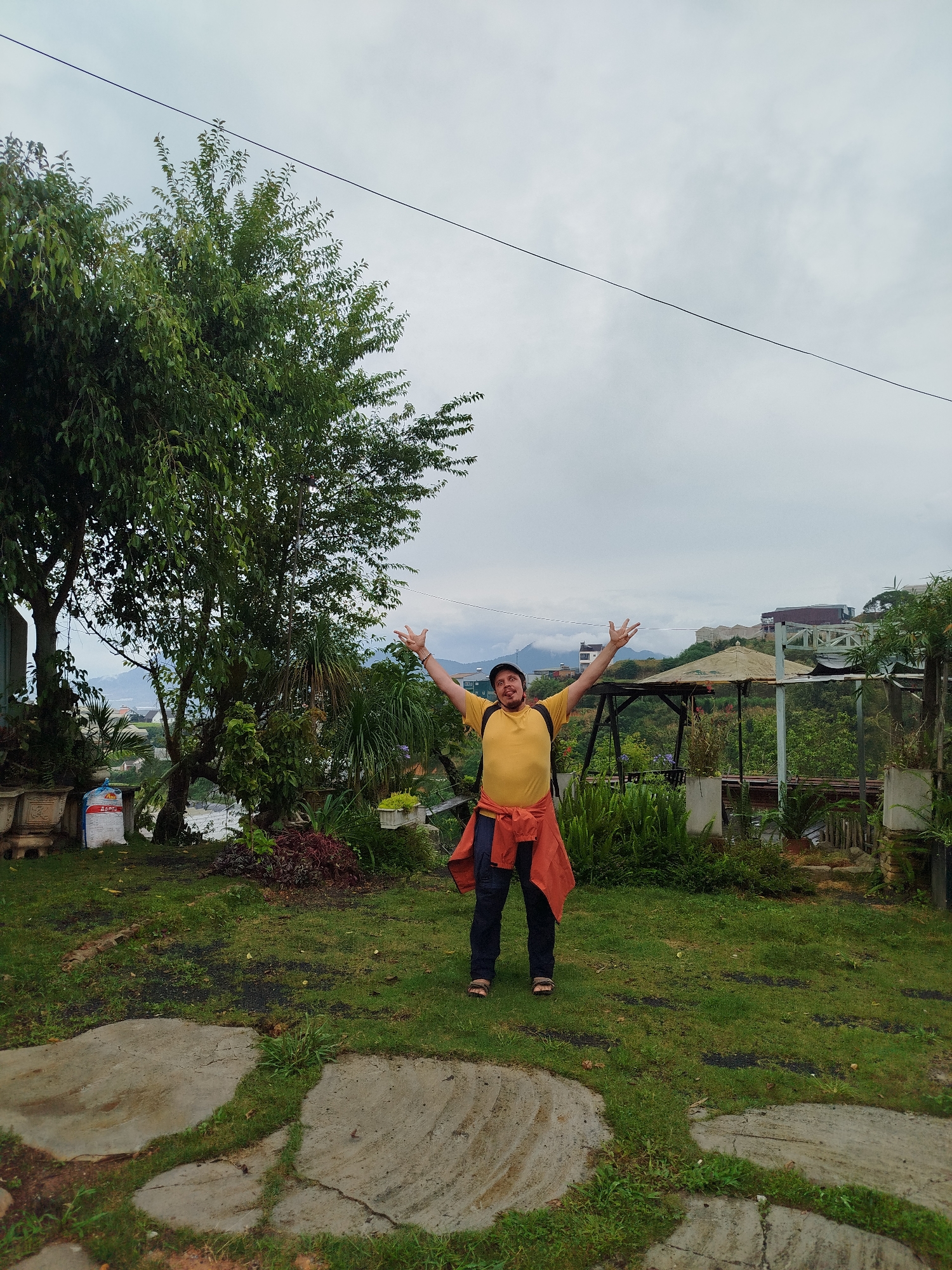 A man stands with his arms raised in a verdant garden, against a backdrop of cloudy sky and distant buildings in Da Lat, Vietnam. He is wearing a yellow collared shirt, dark pants, and sandals, with an orange jacket tied around his waist and a black hat on his head. The garden features lush greenery, including a large tree to the left and various plants scattered around. A stone pathway stretches in the foreground. In the background, there is a structure with a tent-like roof and other buildings nestled on a hillside. An overhead wire spans the sky.