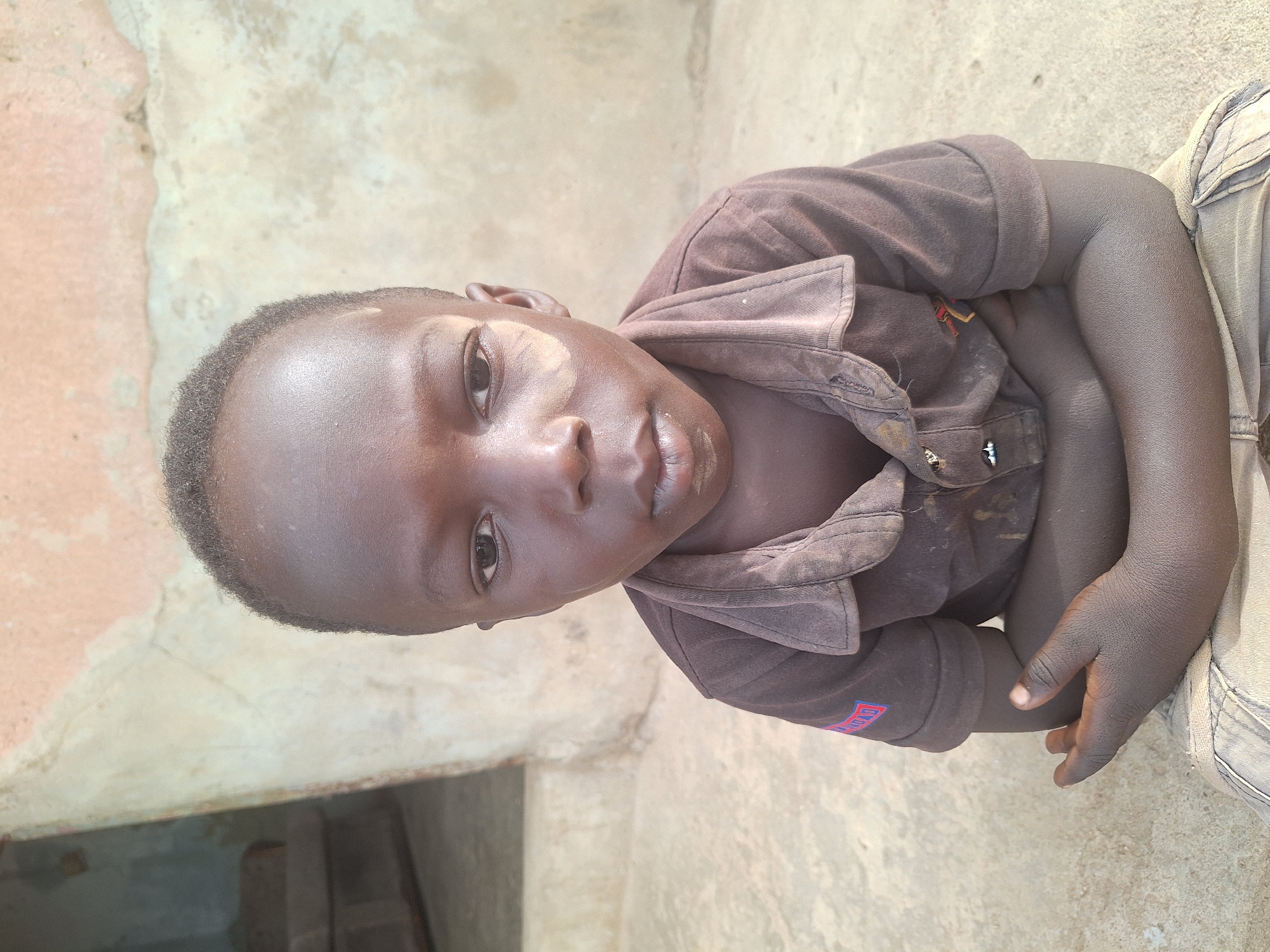 This is a portrait of a young child, likely a boy, with dark skin, captured from the upper chest upwards. The child is looking directly at the camera with a neutral, slightly serious expression. He has short, dark, curly hair.
He is wearing a short-sleeved, collared shirt in a dark brown color, which appears somewhat faded and possibly dusty or worn. The shirt has several buttons down the front. On the left sleeve, a red patch with the word "WORLD" printed in white text is visible. His arms are bent, and his hands are resting, suggesting he is sitting or kneeling. Part of light-colored (possibly beige or grey) trousers or shorts is visible on his lower right side.
The setting appears to be outdoors in daylight. The background consists of rough, light-colored surfaces, possibly concrete or sun-baked earth, consistent with an outdoor environment in Sabon Tasha, Nigeria. The lighting is bright, with some natural highlights on the child's skin.