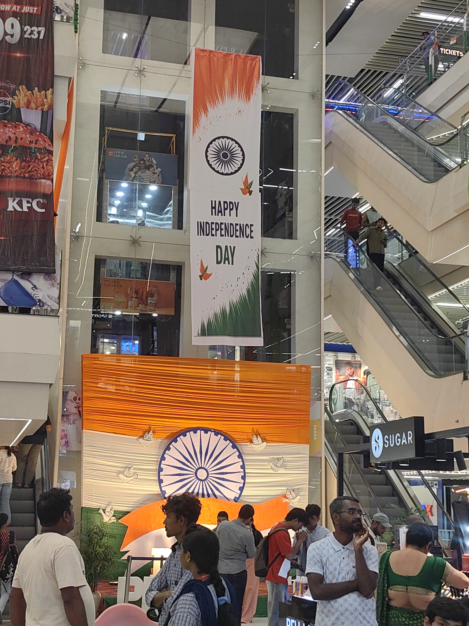 The image depicts an indoor scene, likely a shopping mall, with multiple levels and escalators visible. Dominating the central area is a large banner displaying the Indian flag's colors (saffron, white, and green) with a large Ashoka Chakra in the center. Text on the banner reads "HAPPY INDEPENDENCE DAY." To the left, a large advertisement for KFC featuring a burger and french fries is partially visible.
Several individuals are present throughout the scene. On the escalators, people are seen moving between floors. In the foreground, a group of people are gathered near a decorative display that includes the Indian flag motif. One person is wearing a white t-shirt, another a checkered shirt, and a third a blue uniform. Another individual in the foreground is wearing glasses and a patterned shirt, seemingly engaged in a phone conversation. Further back, more people are visible on escalators and walking around.
The environment appears to be well-lit with artificial lighting. The architecture suggests a modern commercial space with glass railings and tiered structures. There is a sign visible that reads "SUGAR." While the image is taken in Bichhia, India, this specific location within Bichhia is not identifiable from the visual information.