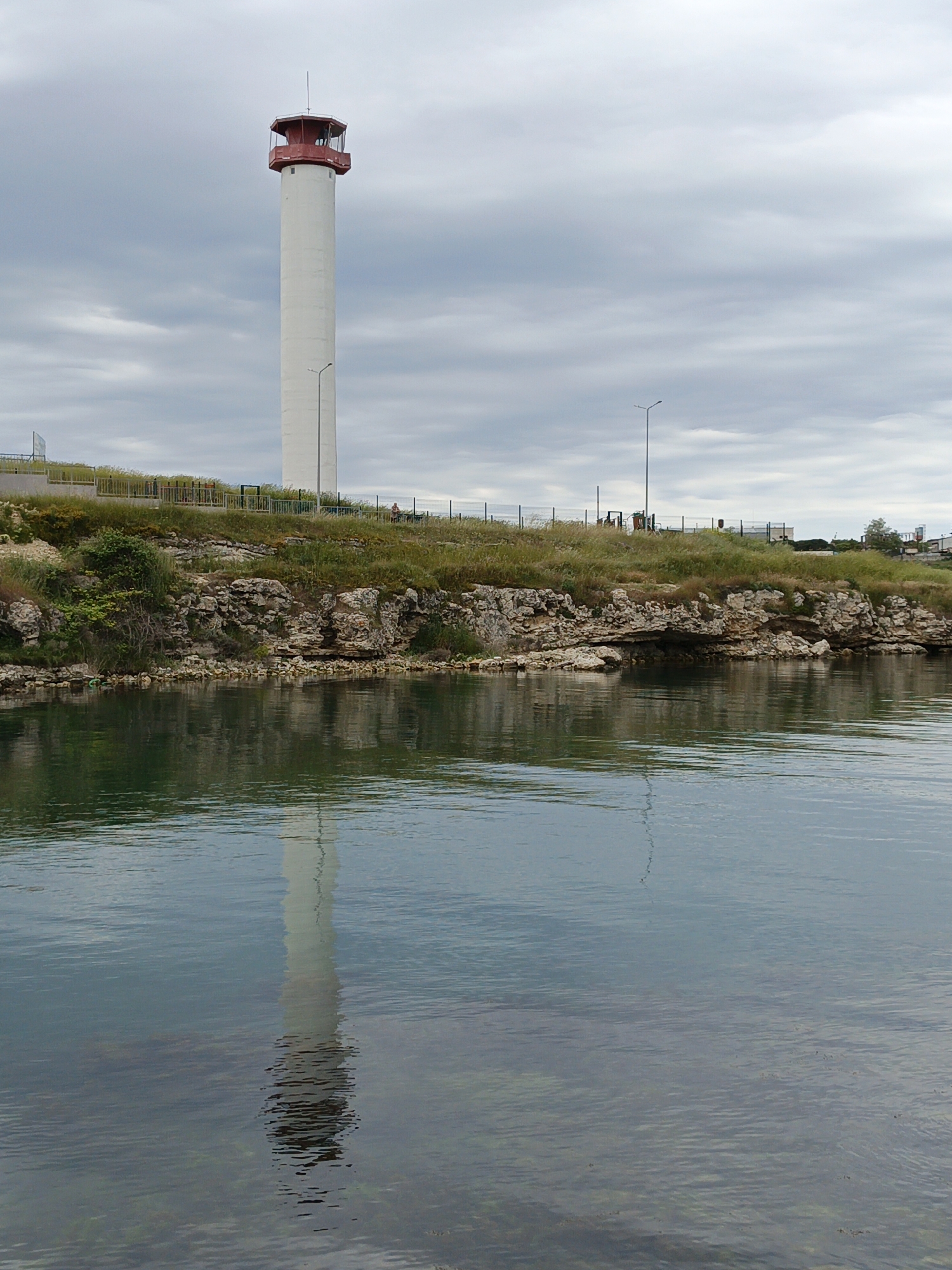 The image shows a tall, white tower with a red-trimmed top standing near the edge of a body of water. The tower appears to be a control tower, with the red trim highlighting the observation area at the top. The tower’s reflection is visible in the water. The shoreline is characterized by rocky terrain, vegetation, and a fence. The sky is overcast. The location is Unknown, Unknown.