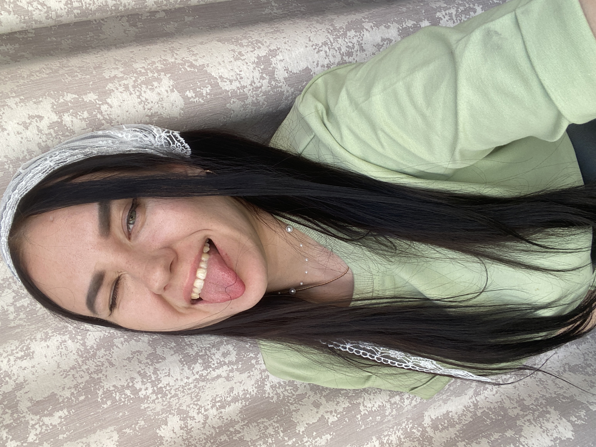 A young woman with long, dark hair and fair skin is lying down on a patterned surface, possibly a bed or sofa. She is wearing a light green top and a white lace headband. Her mouth is open, tongue extended, and she is winking with her right eye. A delicate necklace adorns her neck. The location is Chaikovsky, Russia.