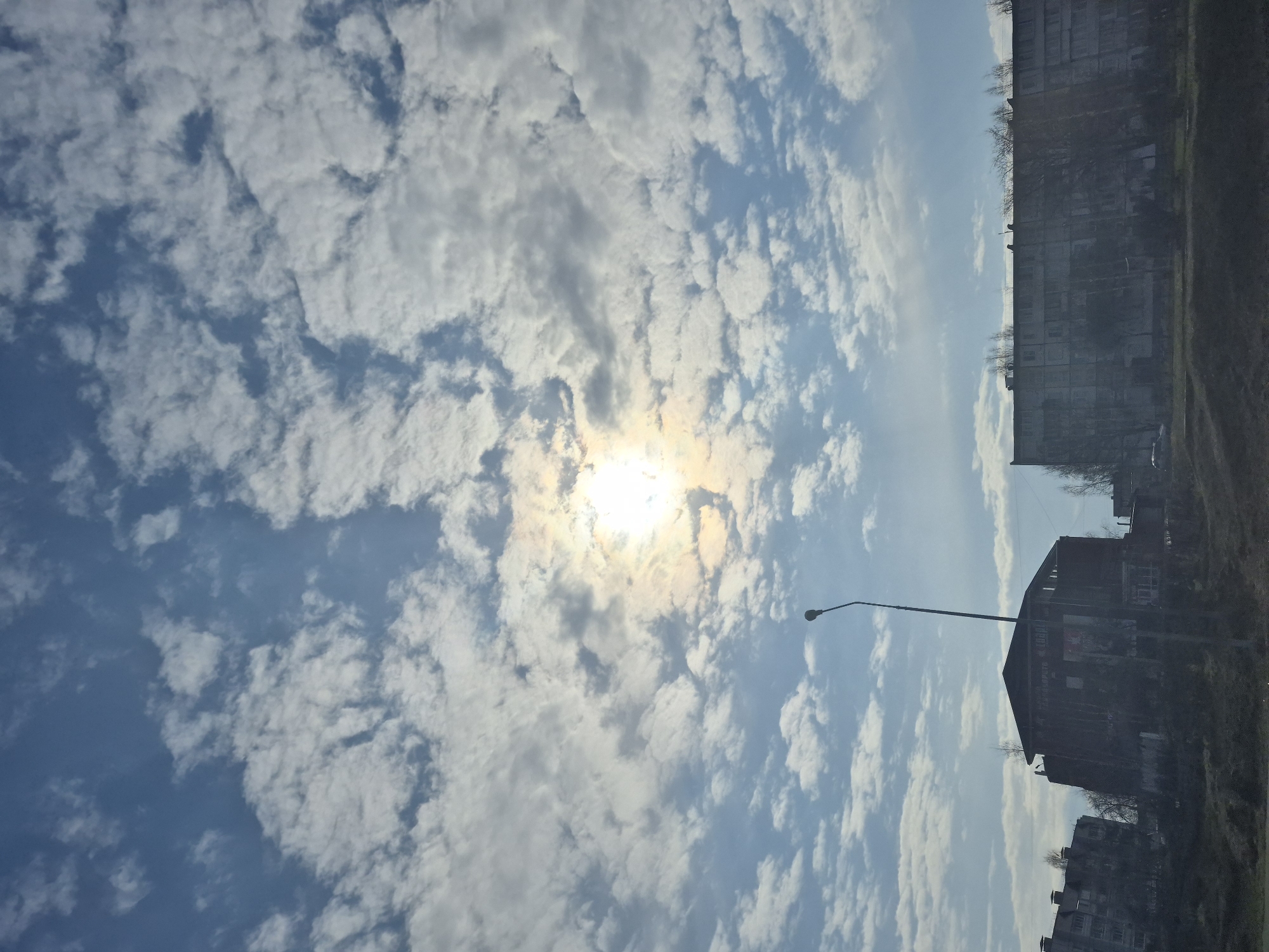 The image depicts a daytime scene in the city of Chaikovsky, Russia. The sky is predominantly blue with extensive cloud cover, and the sun is visible, partially obscured by clouds. In the lower-right portion of the image, there are silhouettes of buildings. A lamppost extends towards the clouds.
