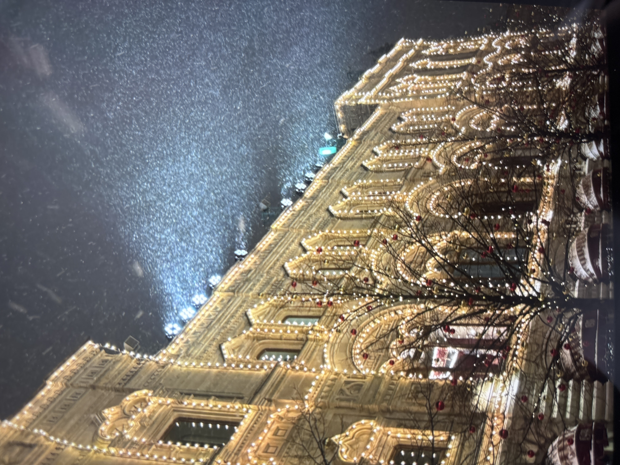 At night, the facade of a large building is brightly illuminated with numerous lights, appearing golden. Trees with red ornaments are in front of the building, obscuring part of it. It is snowing heavily. The location is a city, possibly the GUM department store in Moscow.