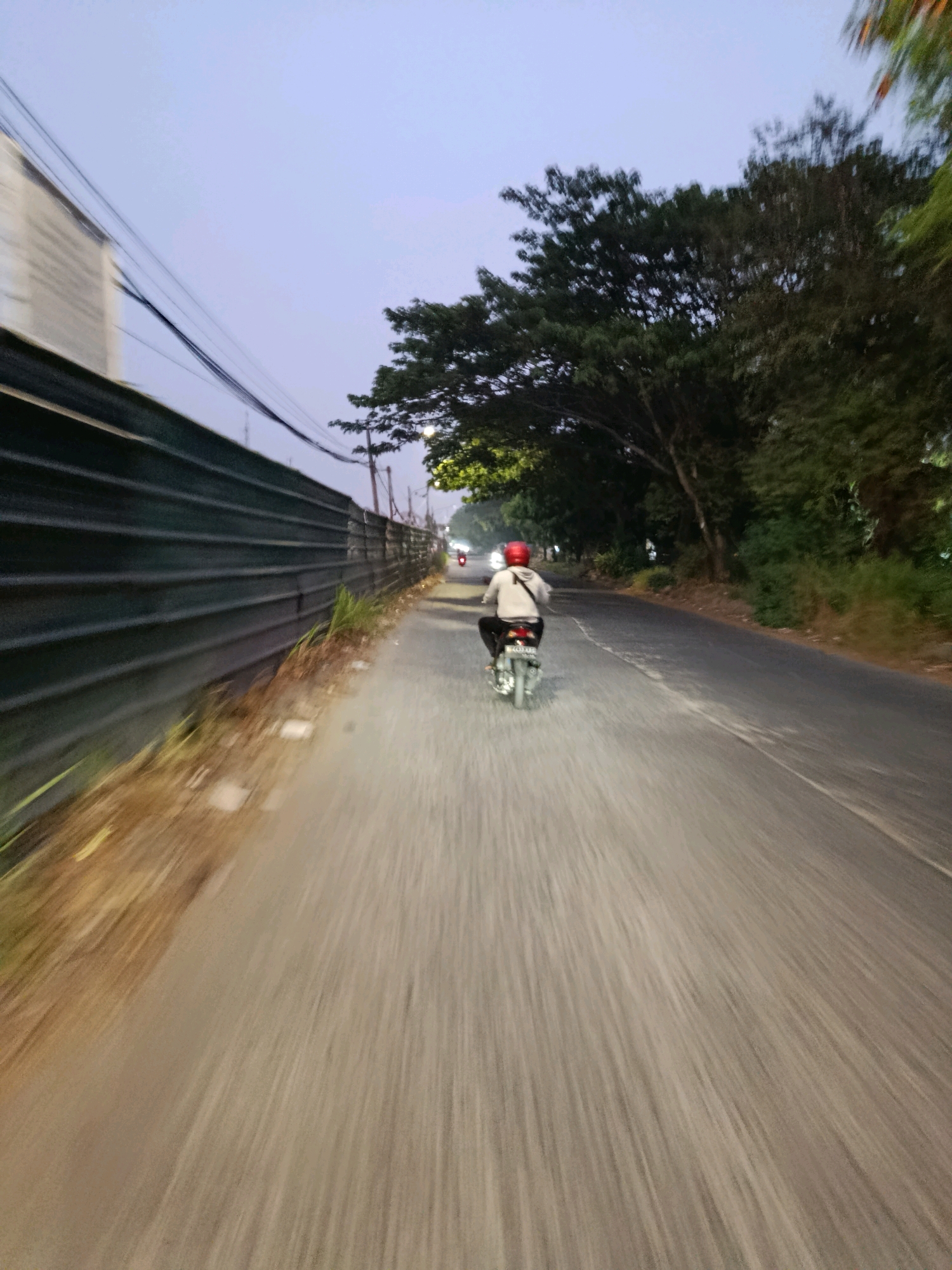 A person rides a motorcycle on a road in Bekasi, Indonesia, with a dark fence and trees lining the left side of the road. The sky is a pale blue, and the time of day appears to be dusk or early morning. The motorcycle is silver, and the rider is wearing a white hoodie and a red helmet. The road is asphalt, and there is a slight blur from the motion of the motorcycle, suggesting it is moving at a moderate speed. The fence appears to be made of corrugated metal, and there are some lights visible in the distance along the road.