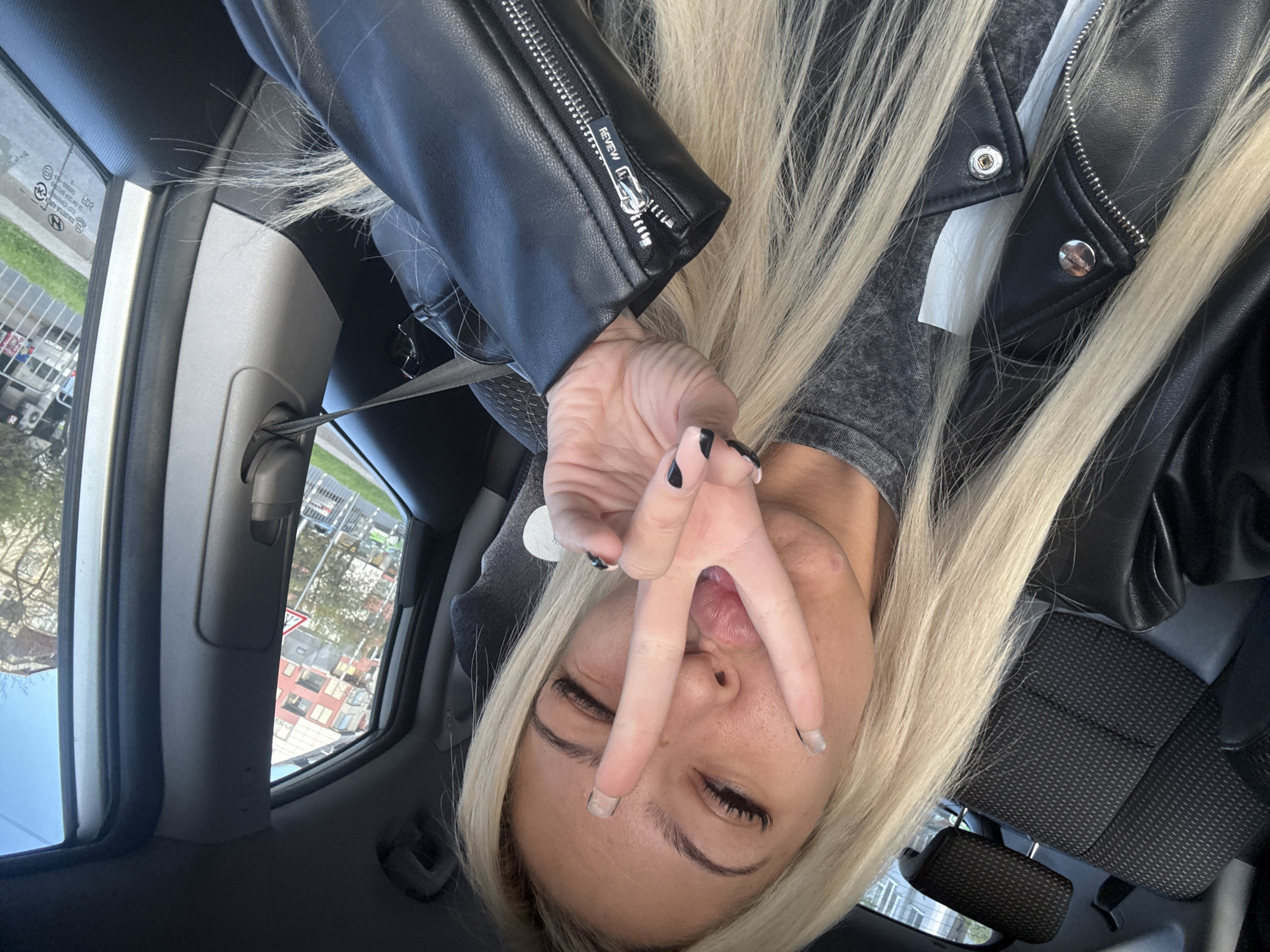 A woman with long, blonde hair and wearing a black leather jacket is sitting inside a vehicle. She is making a hand gesture with her fingers spread, partially covering her face. The vehicle's interior includes a gray dashboard, a seat with a textured black fabric, and a view of the city outside through the window. The city appears to have buildings and greenery. The woman is wearing a seatbelt. The location in the city is unknown.