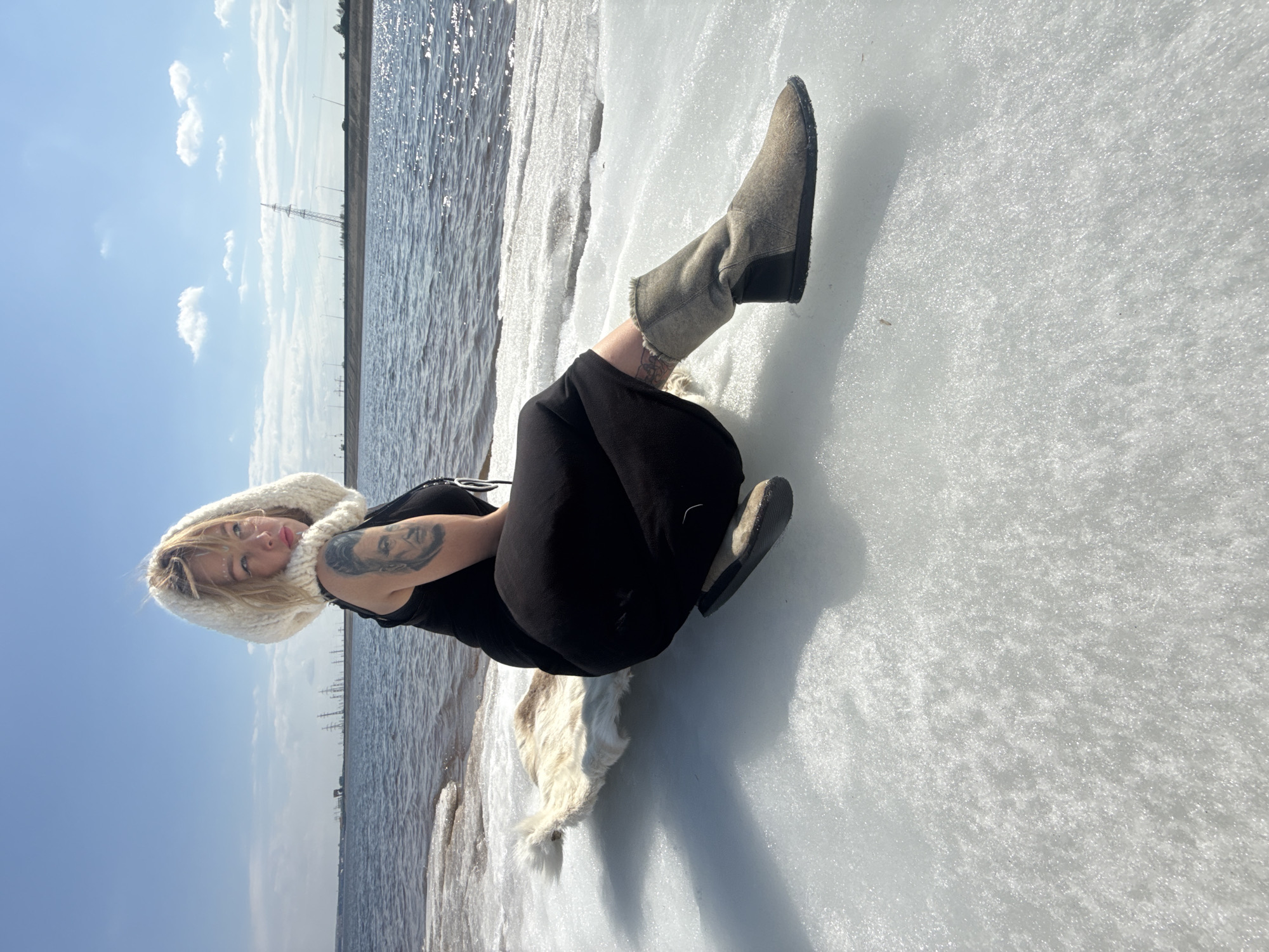 The image, viewed in correct orientation, captures a young woman posing outdoors in Noviy, Russia, on a bright, clear day. She is lying on her side on a vast expanse of sparkly white ice or snow, facing slightly towards the right and looking directly at the camera with a serene expression. She is dressed in a black sleeveless top, black pants, and a cozy, cream-colored fluffy hood. Her feet are clad in gray, mid-calf boots. Tattoos are visible on her left upper arm and lower leg. Adjacent to the ice, a body of water with rippling waves catches the sunlight, glistening brightly. The background reveals a distant shoreline or embankment, featuring utility poles and a prominent electrical tower under a clear blue sky with scattered white clouds. The sun intensely reflects off both the water and the shimmering texture of the ice. A piece of white fur or fabric rests near her hip. The scene conveys a tranquil moment in a cold, sunny, and natural environment.